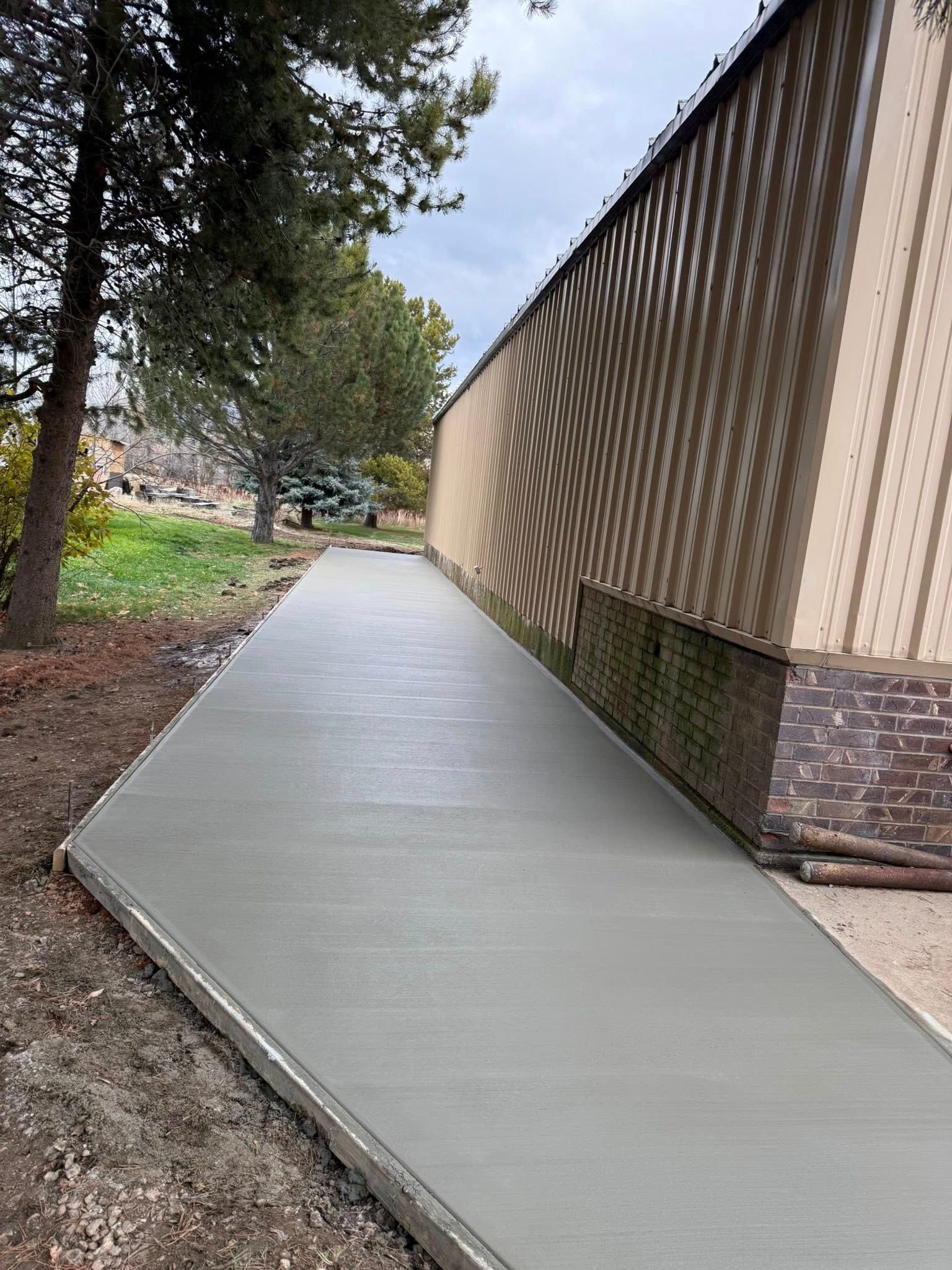 A newly poured gray concrete walkway stretches along the side of a building next to a treed area.