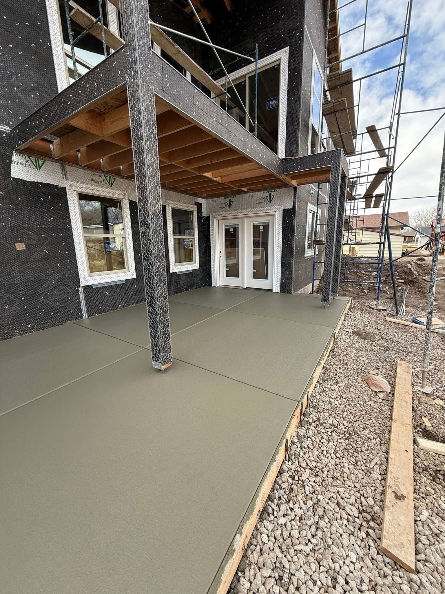 A newly poured concrete patio attached to a house under construction, featuring a support pillar and gravel ground.
