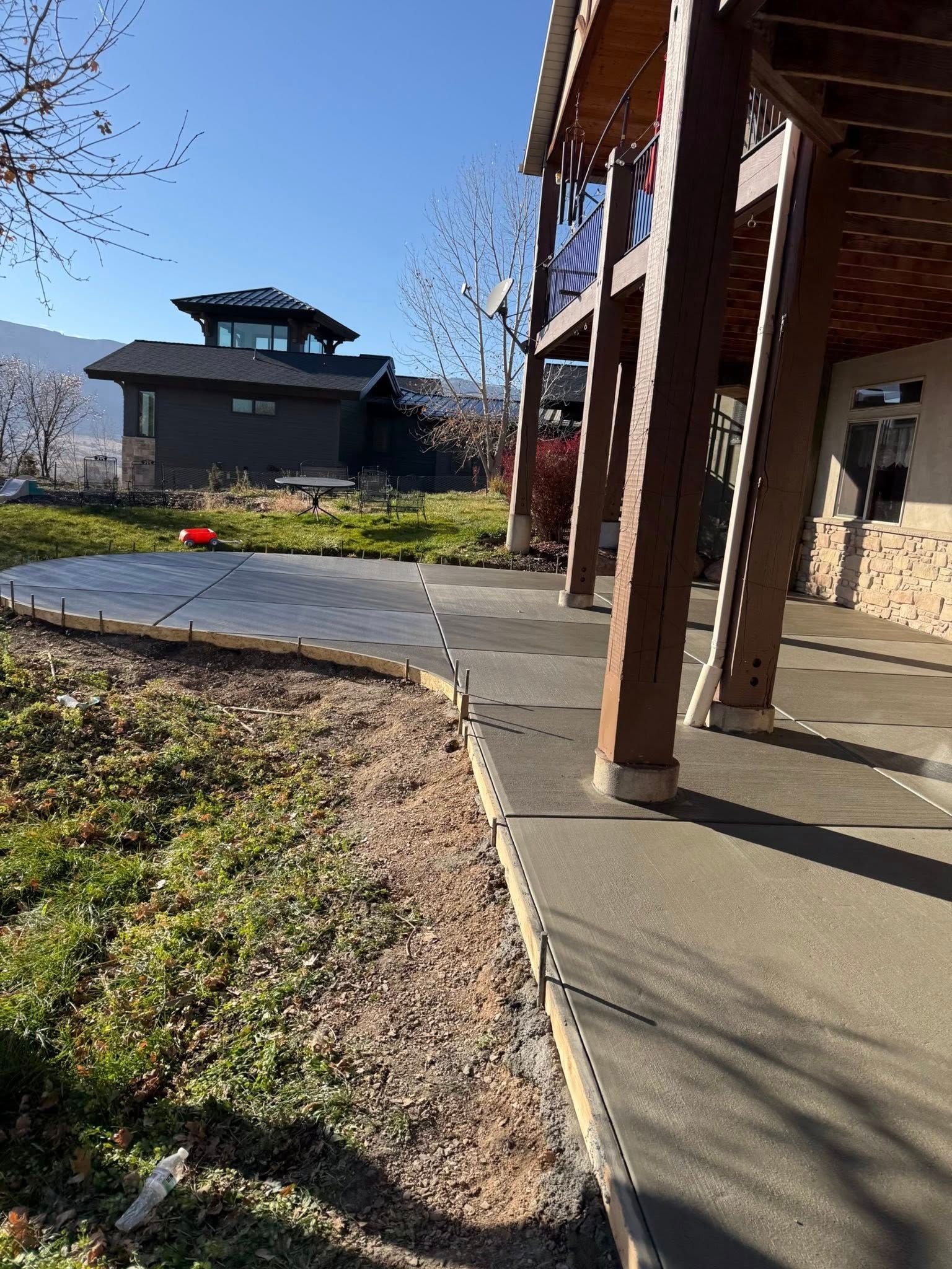 A new concrete patio extends from under a wooden deck toward a grassy lawn and a distant house under a blue sky.