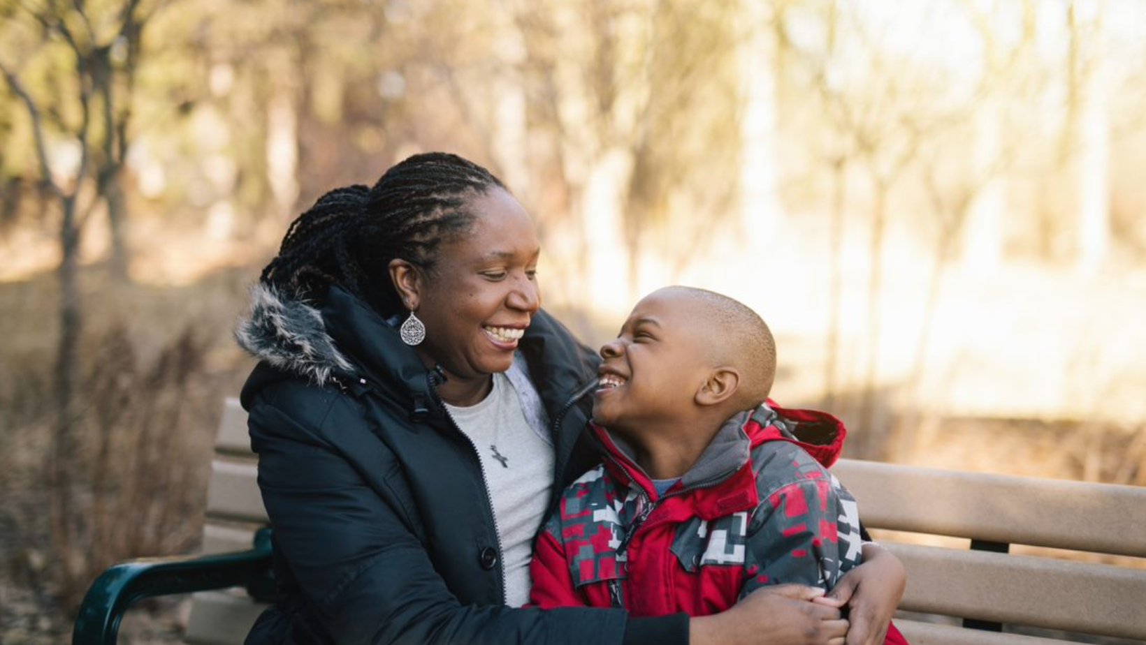 A woman is sitting on a park bench hugging a young boy.