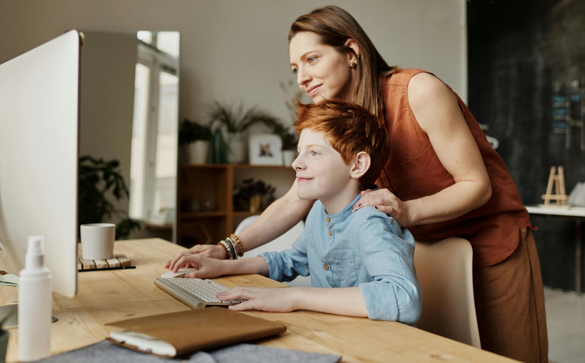 A woman is standing next to a boy sitting at a desk using a computer.