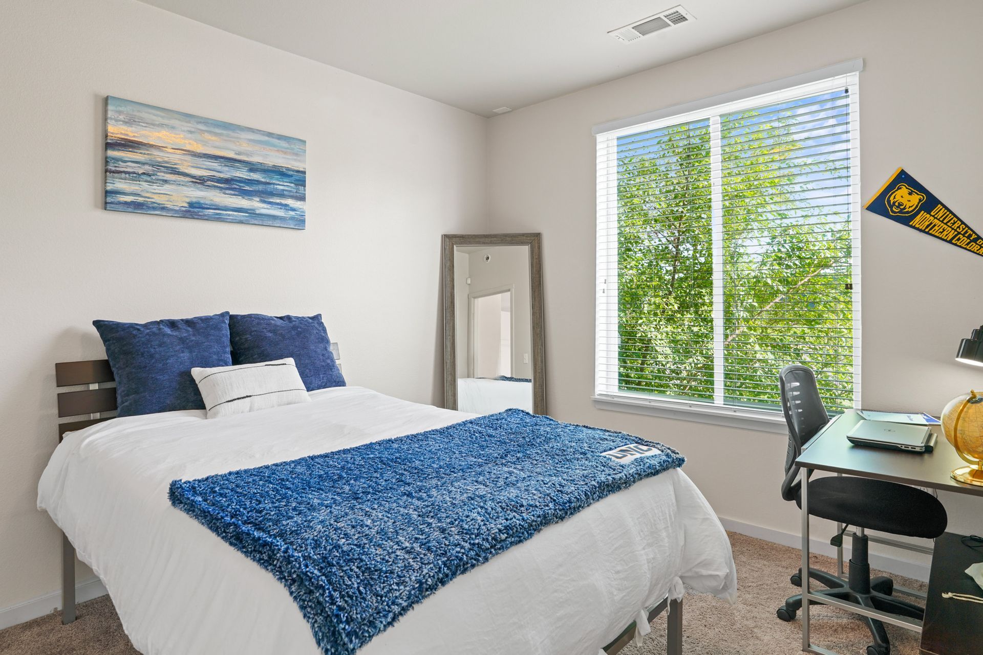 Bright bedroom with a white bed, blue bedding, desk by a window, and a full-length mirror
