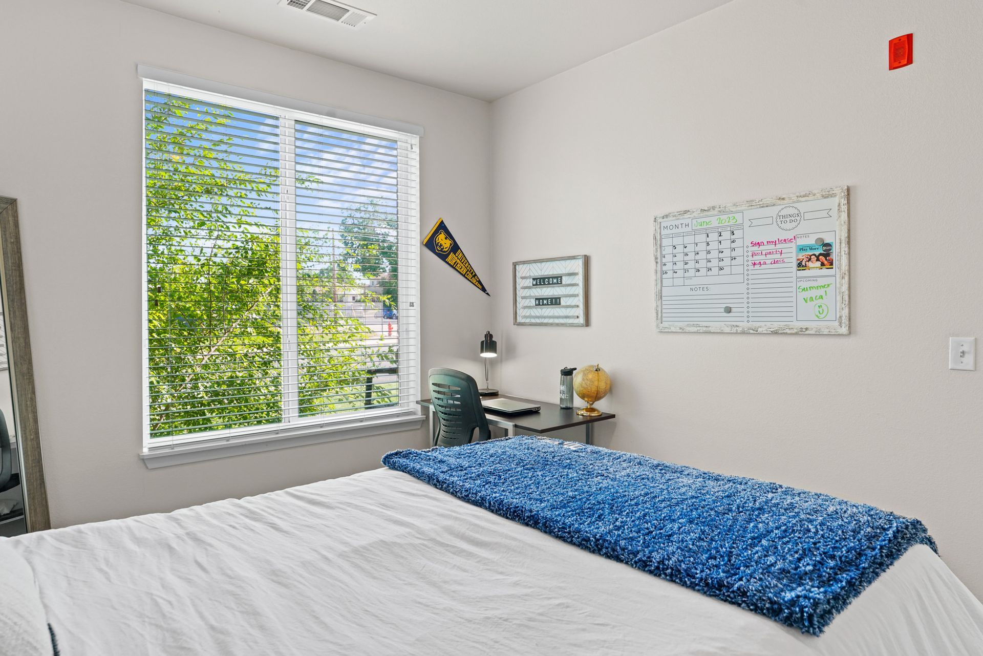Bright bedroom with a bed, blue blanket, desk, window with blinds, and wall decor