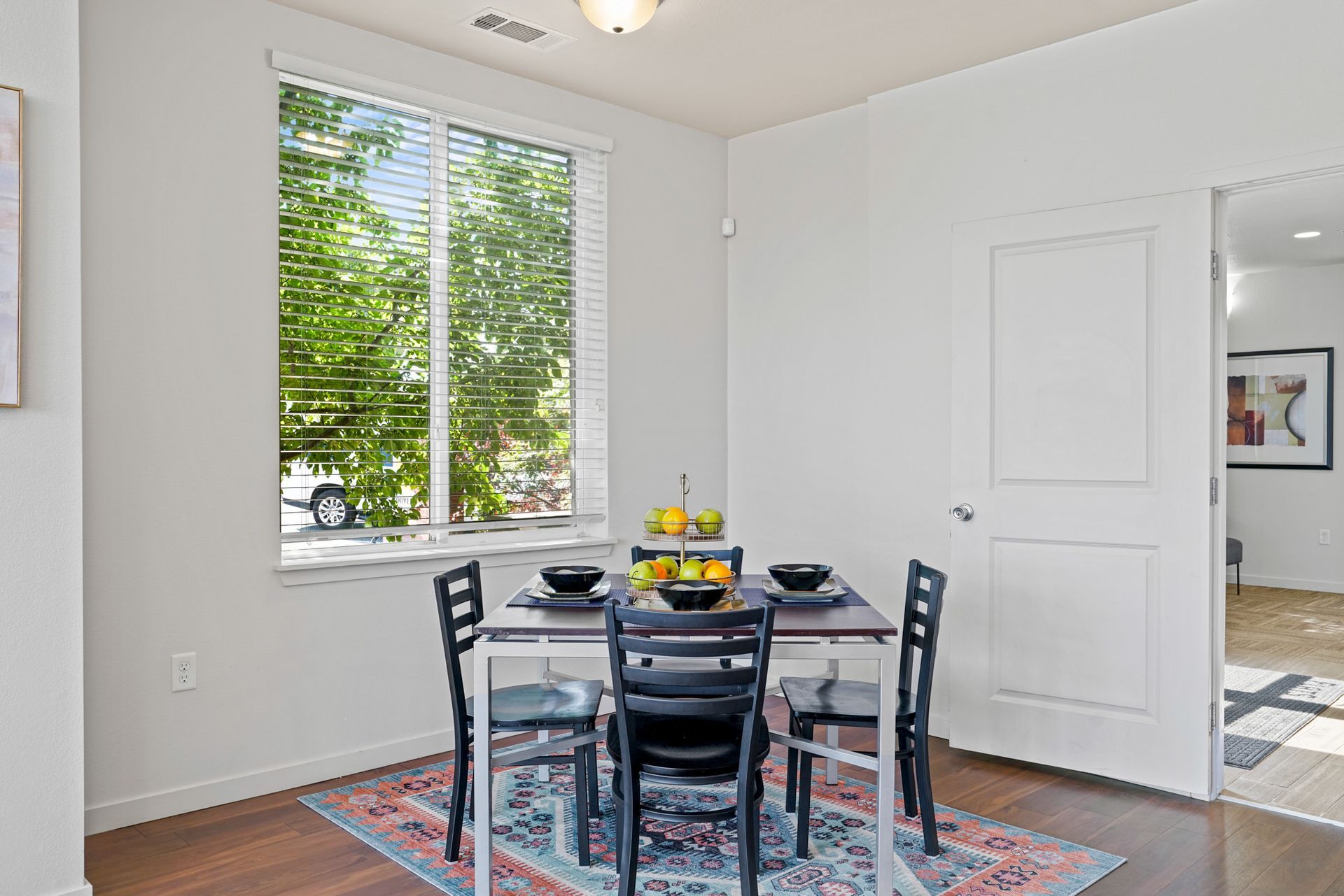 Bright dining nook with black table and chairs, colorful rug, and large window with green trees outside