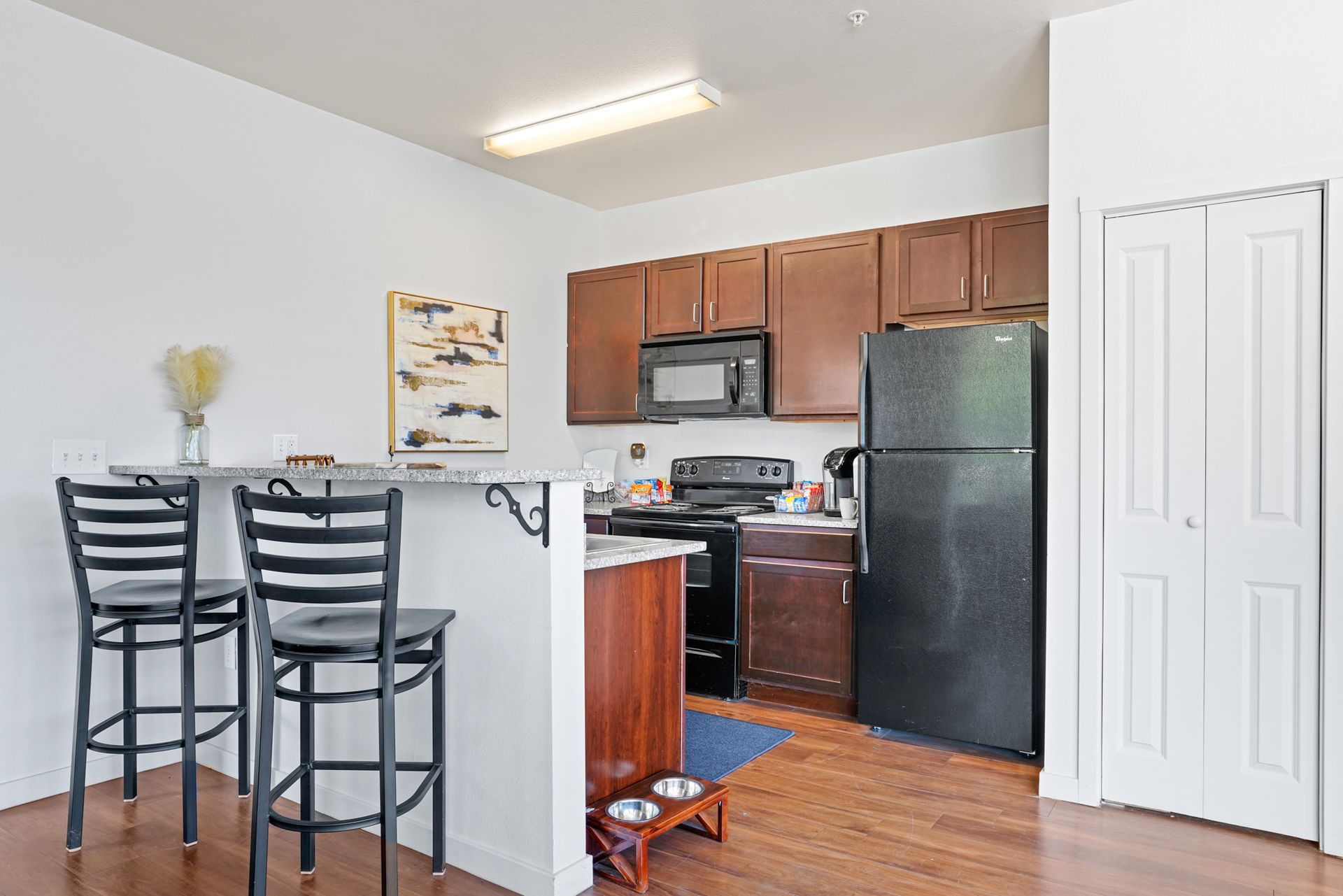 Small apartment kitchen with black appliances, wood cabinets, and a breakfast bar with two stools.