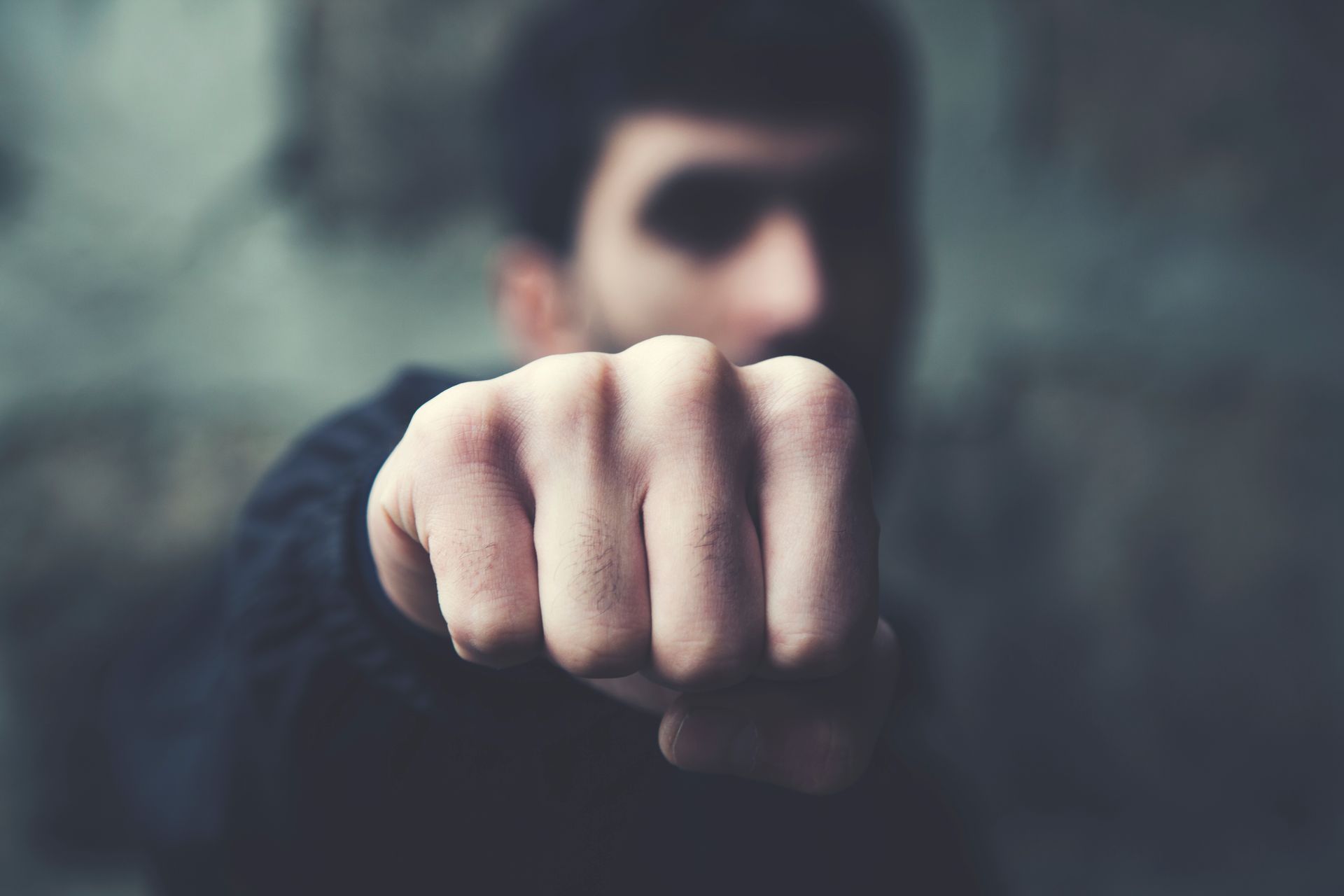 Man's clenched fist thrust forward, blurred face in background, dark setting.
