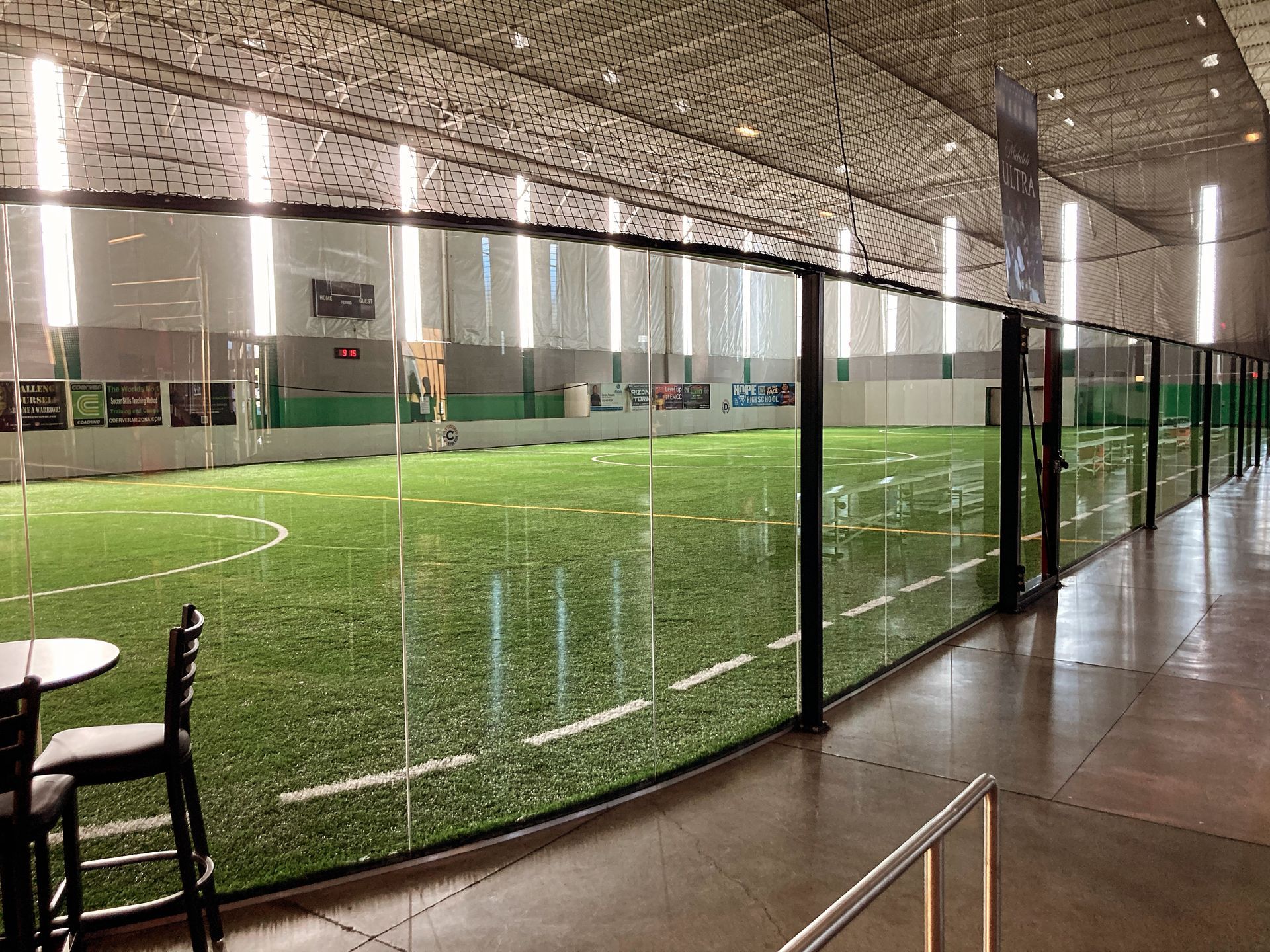 Indoor sports facility featuring a turf soccer field behind a glass wall, viewed from a spectator seating area.