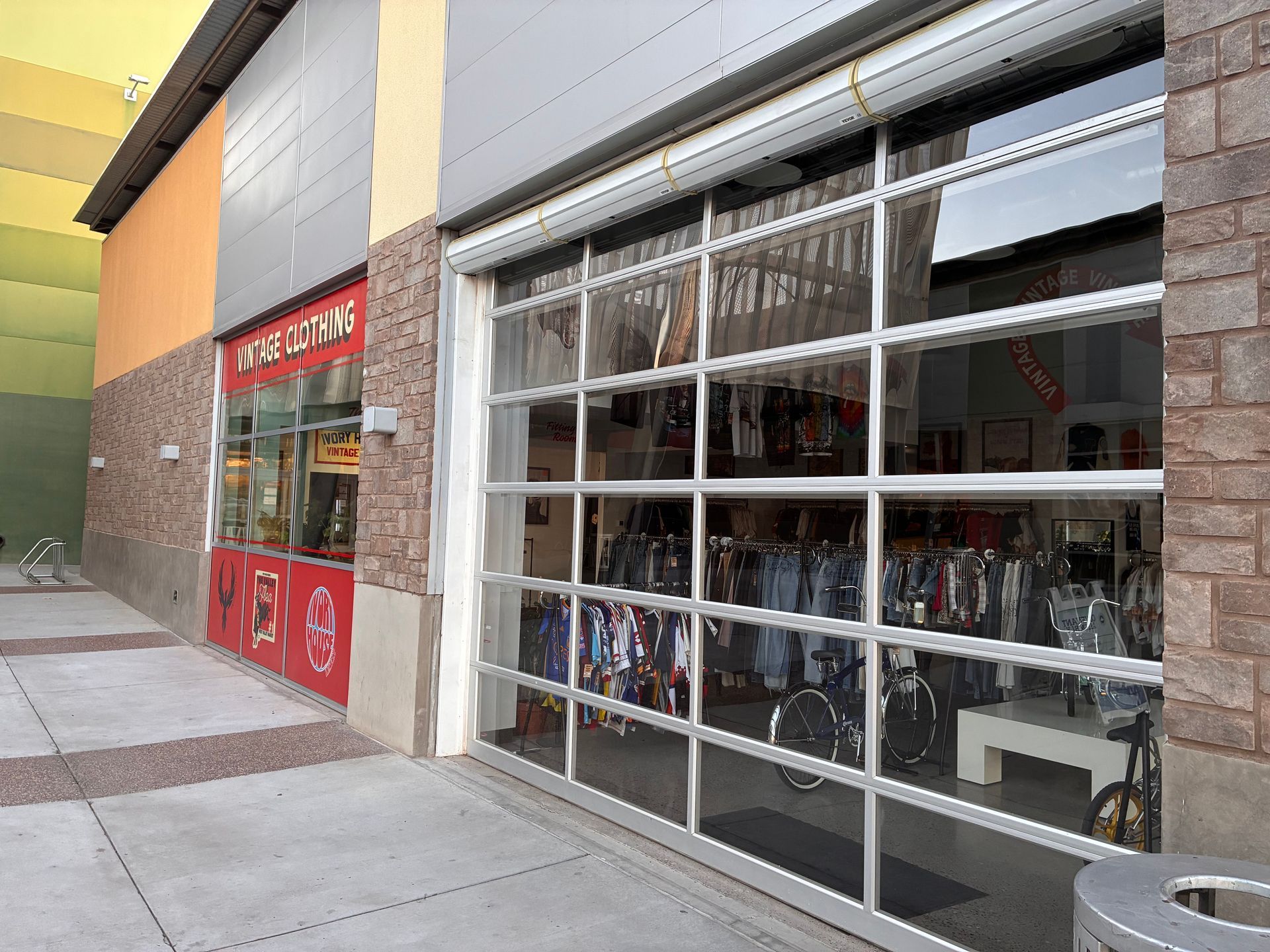 Storefront with a large glass garage door open to a retail space filled with clothing on racks, set in a stone-faced plaza.