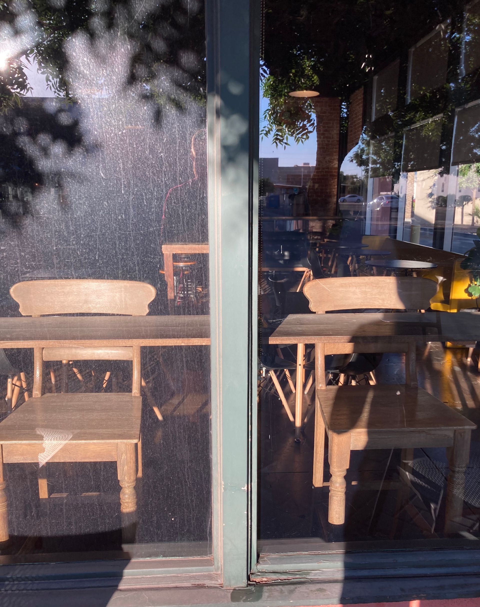 Two wooden chairs and tables seen through a glass storefront, reflecting trees and a bright sunny street.