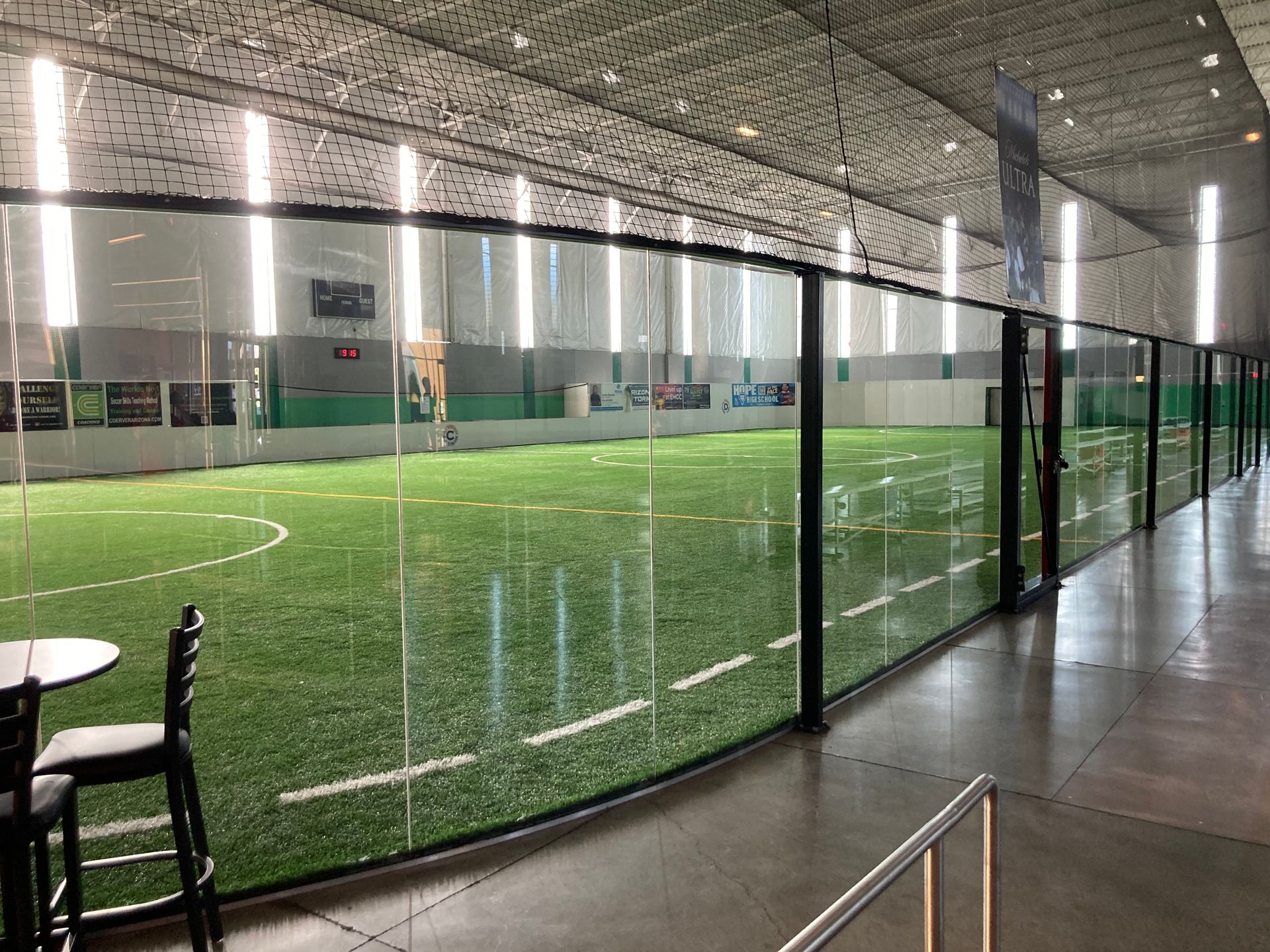 An indoor soccer field with artificial turf, viewed through a glass partition from a modern lounge area with chairs.