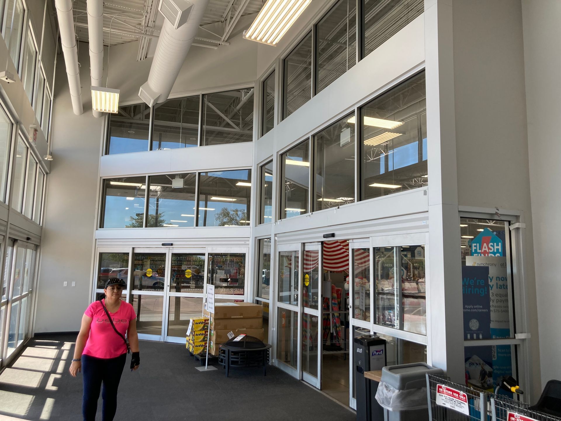 A person in a pink shirt walking through the bright, glass-walled entrance of a large retail store.