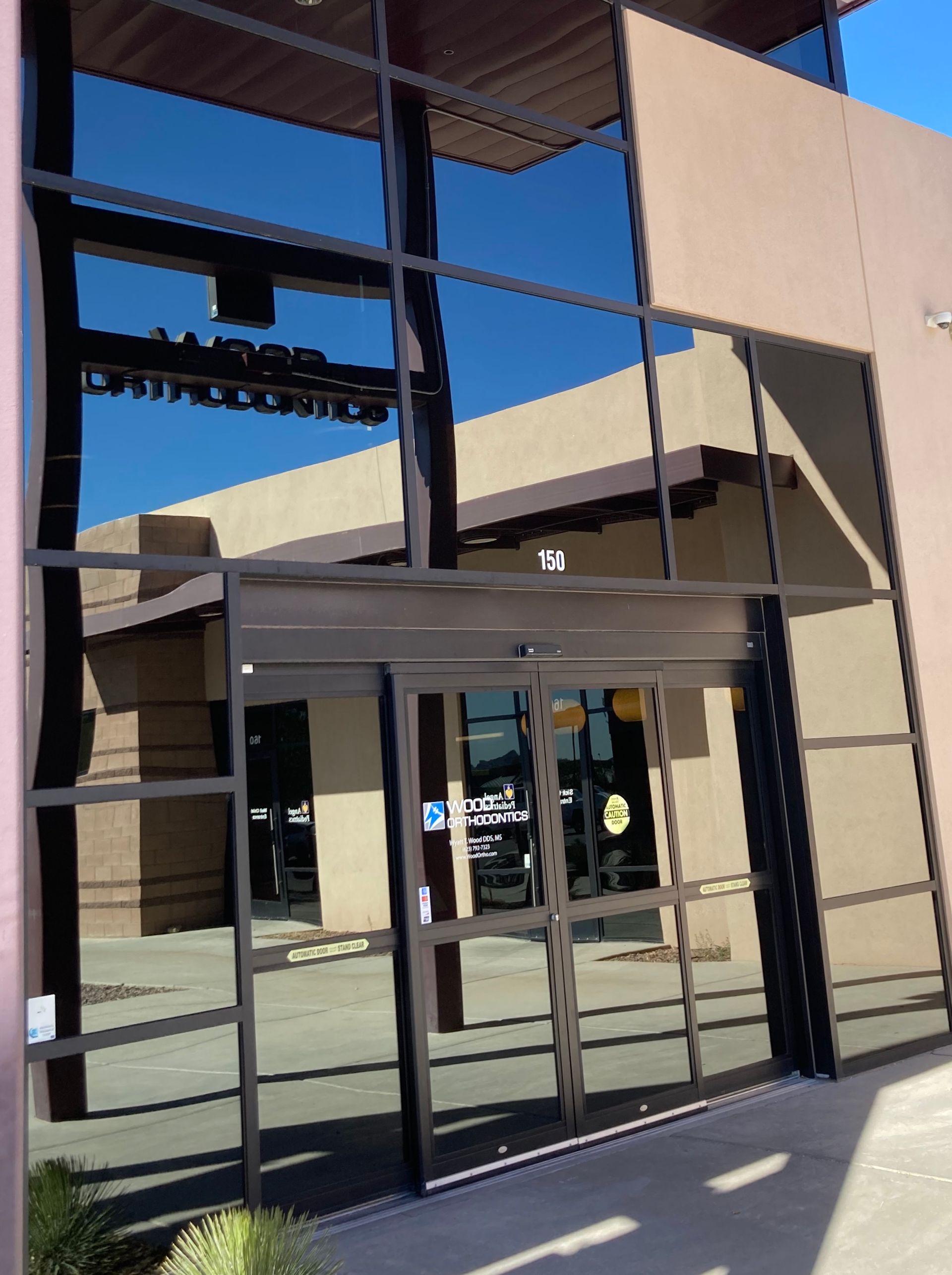Modern glass building entrance with automatic sliding doors and tan exterior walls under a bright blue sky.
