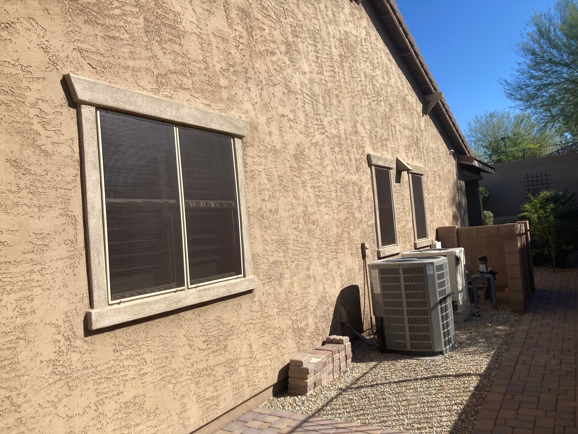 Tan stucco house exterior with a dark-screened window and HVAC units sitting on gravel beside a brick walkway.