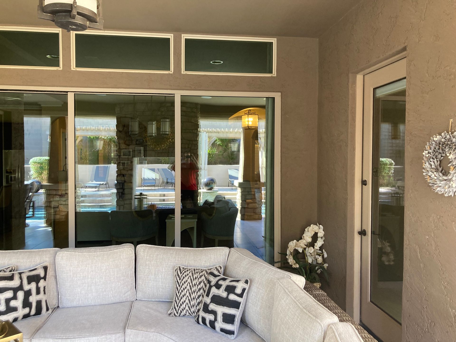 A covered outdoor patio featuring a light gray sectional sofa with patterned pillows, facing a large glass window wall.