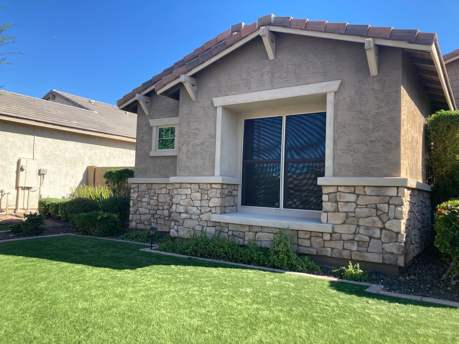 Single-story house exterior with tan stucco, stone veneer accents, a dark-screened window, and a green lawn under blue sky.