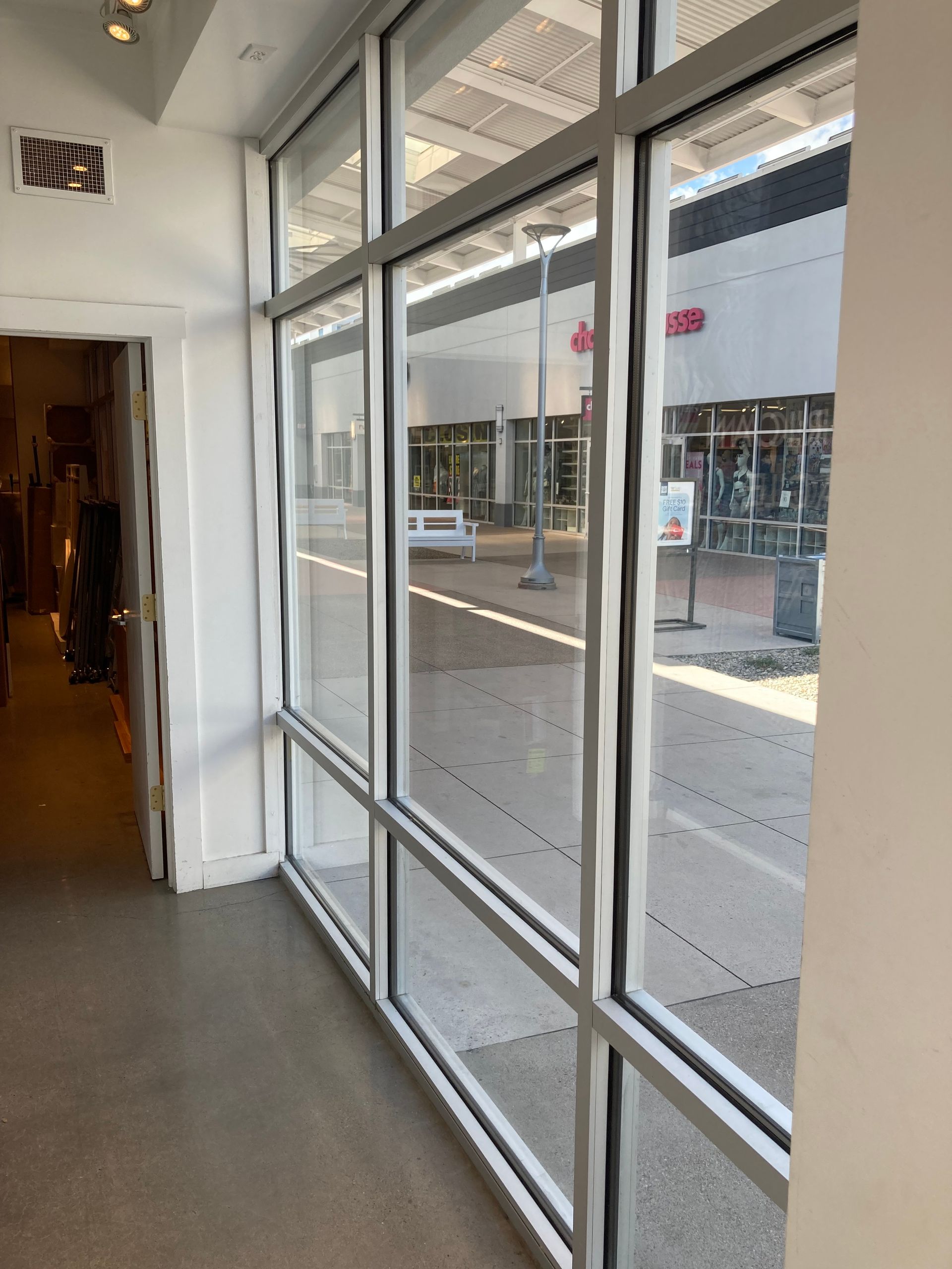 Interior view through large floor-to-ceiling windows looking out onto an outdoor pedestrian walkway in a shopping plaza.