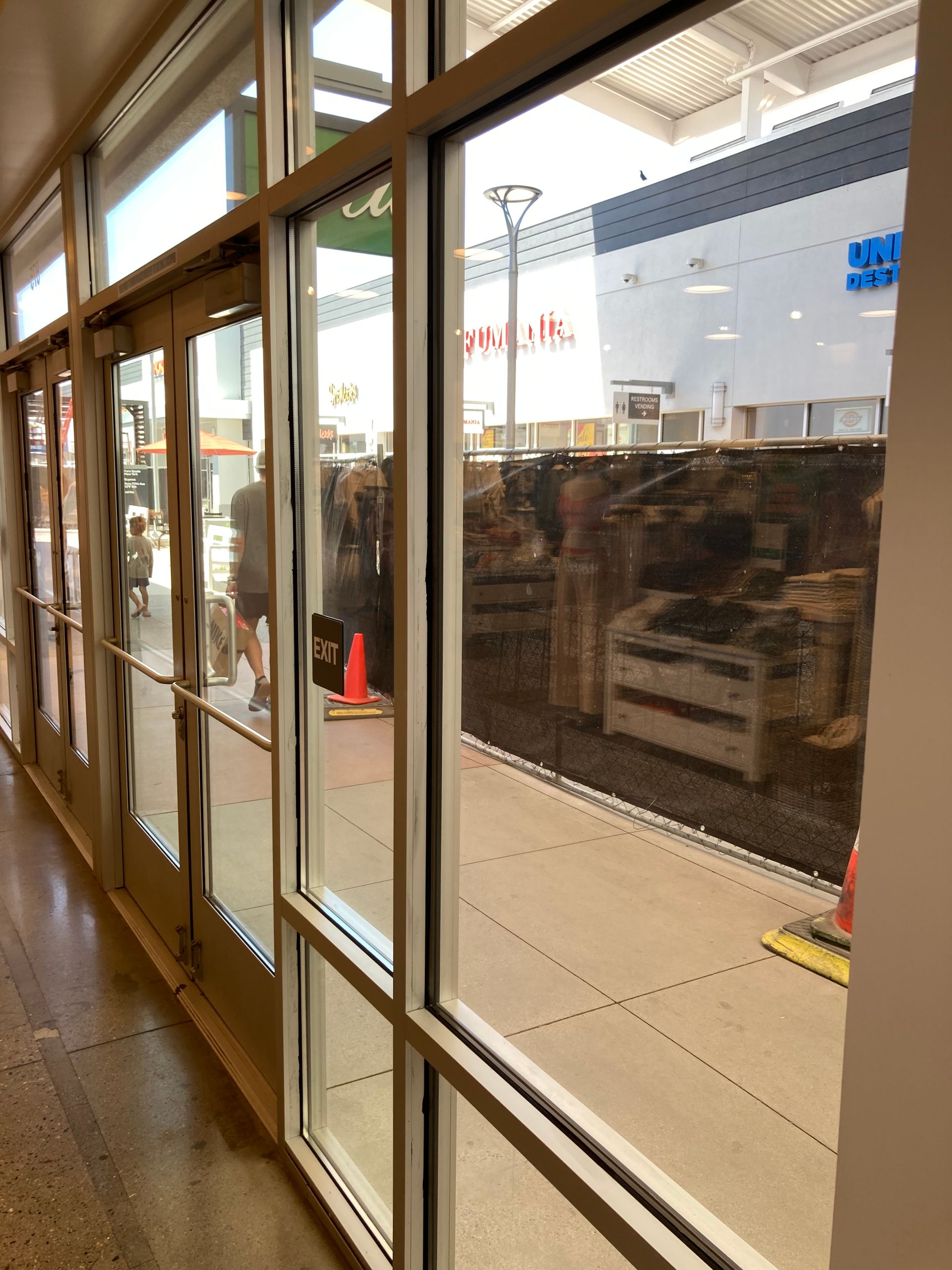 A view through large glass doors showing a person walking past a building with construction fencing and traffic cones.