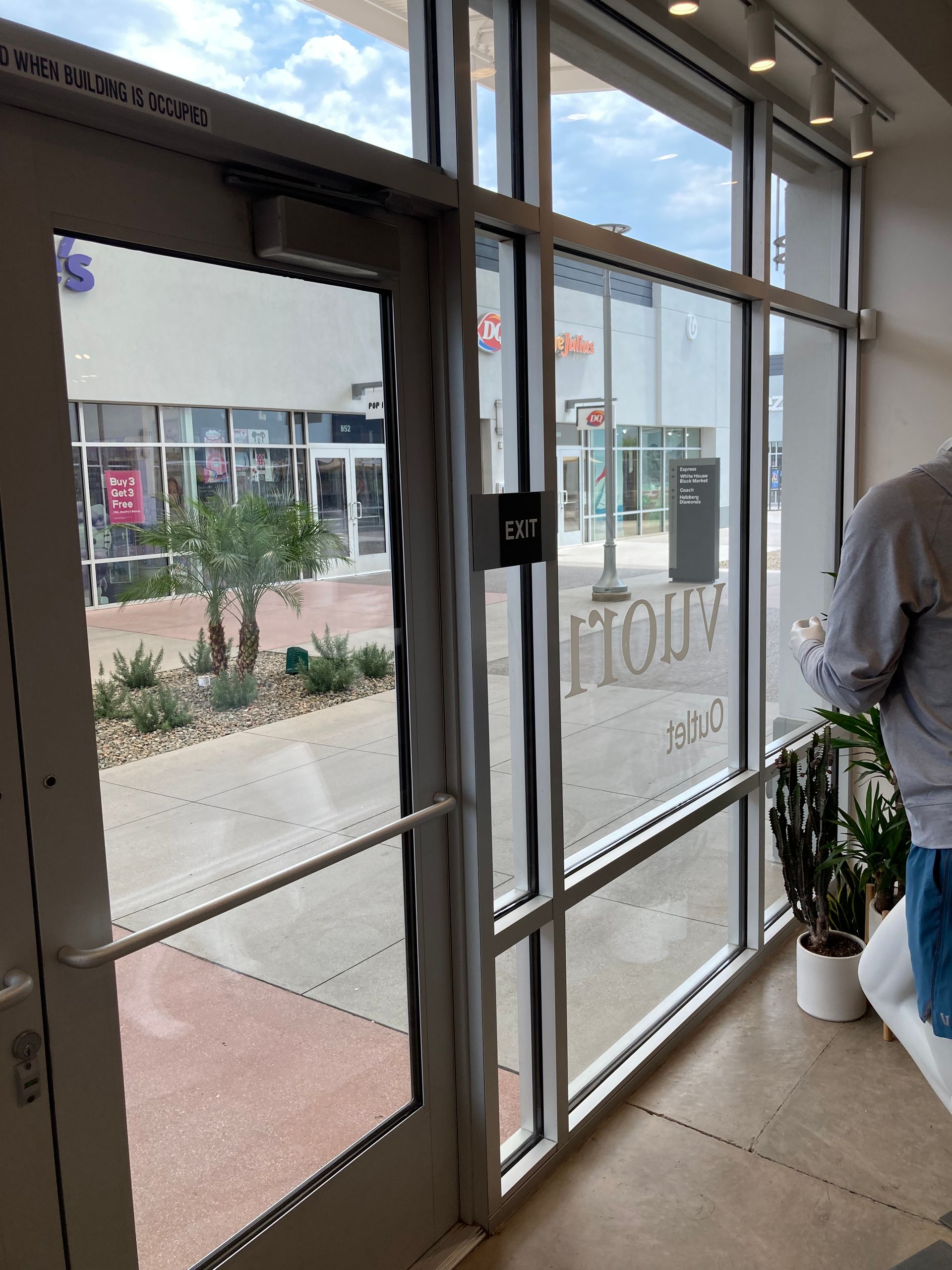 View through a glass storefront door looking out onto an outdoor shopping center plaza with a potted plant inside.