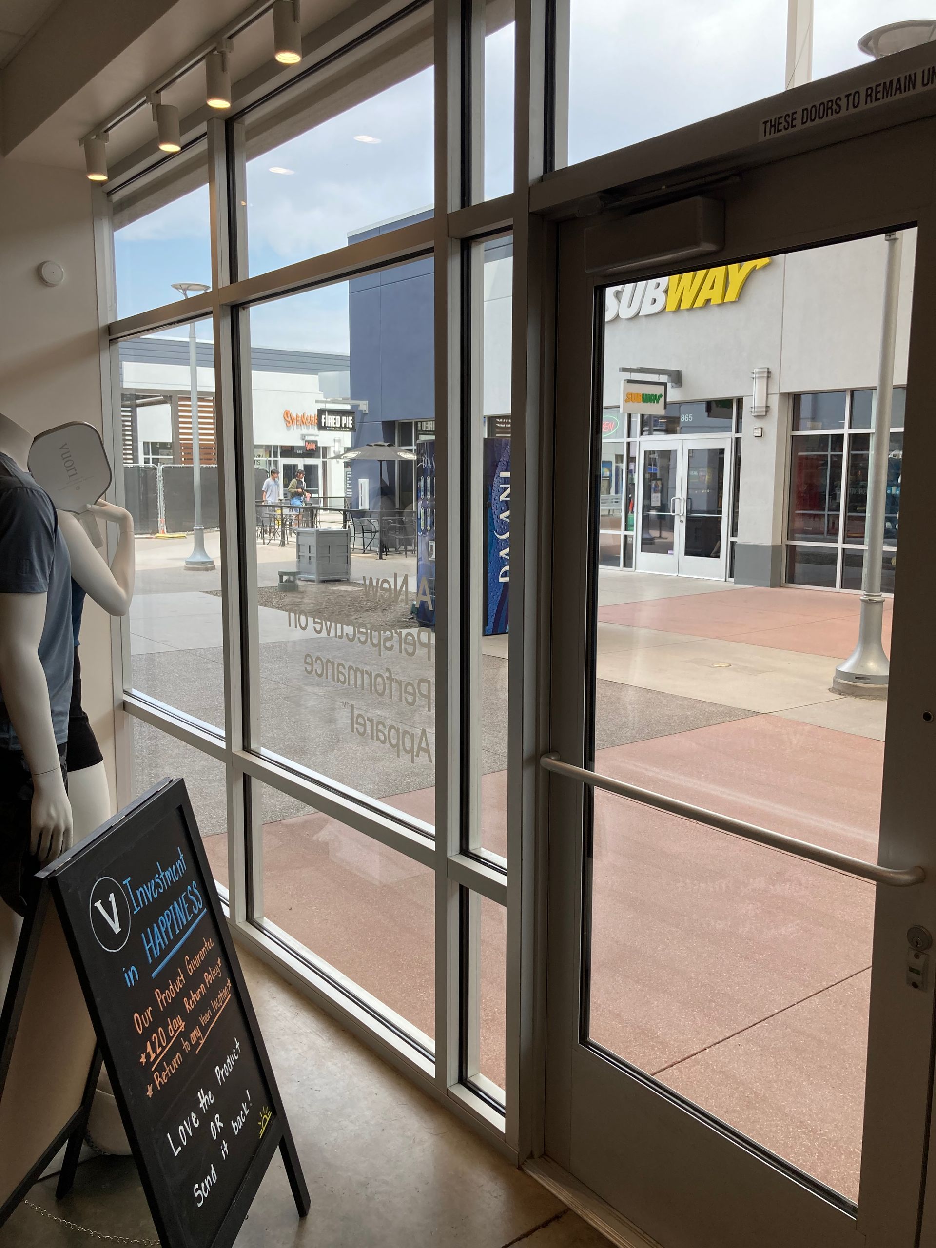 View from inside a retail store looking out a glass door and window at a paved walkway and a Subway restaurant storefront.
