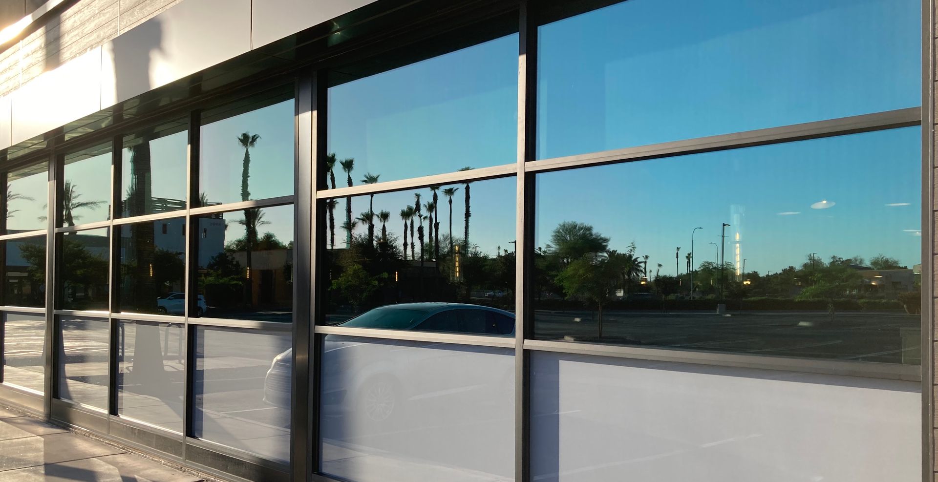 A close-up of a modern building facade with blue-tinted windows reflecting a parking lot, trees, and the blue sky.