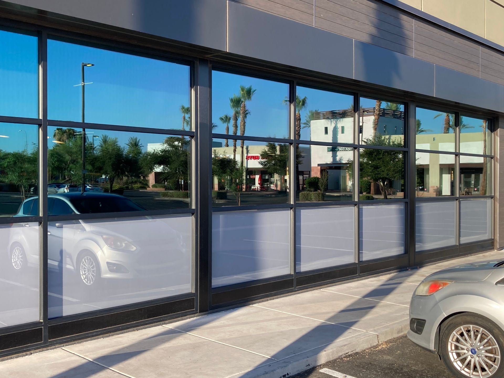 A silver car parked in front of a modern building with blue-tinted windows reflecting a street scene with palm trees.