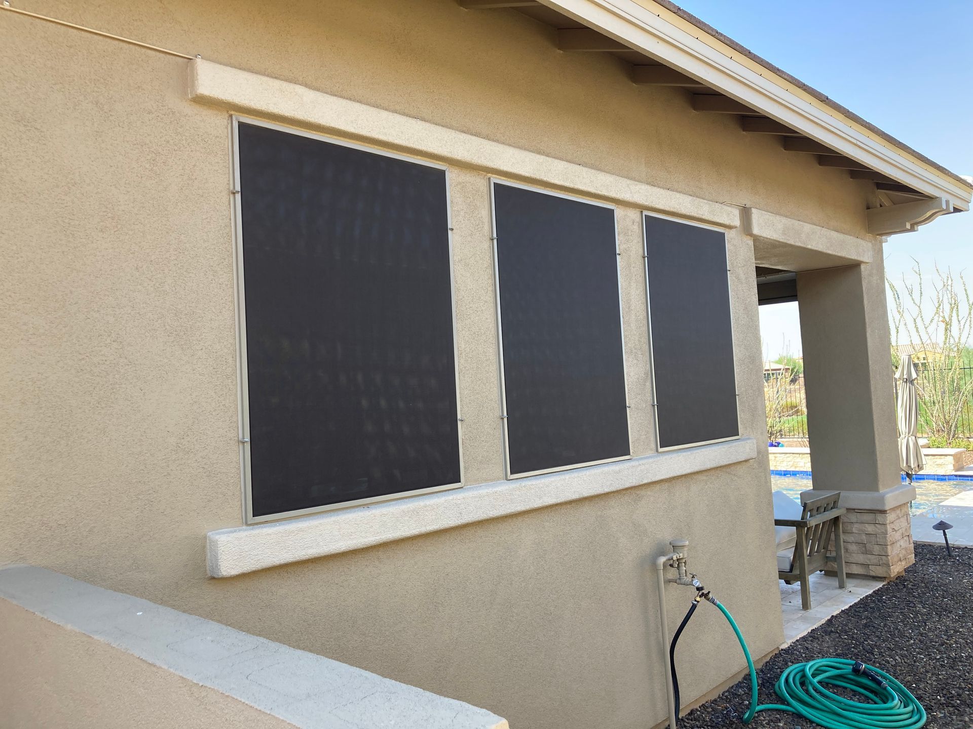 Three rectangular, black exterior solar window screens mounted on the side of a tan stucco house with a porch.