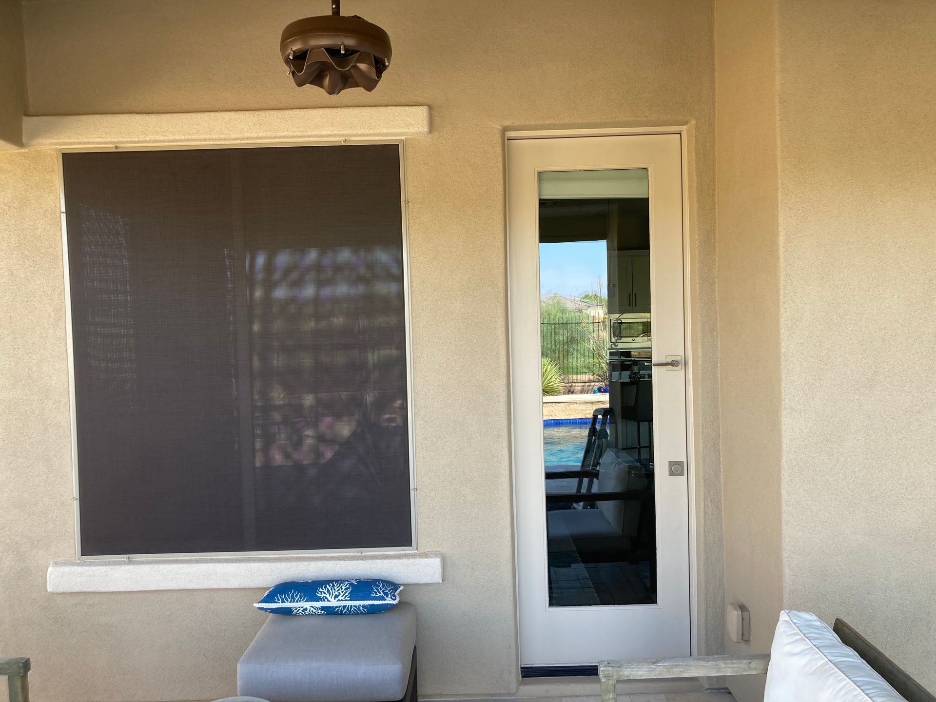 A beige stucco exterior wall with a dark sun-shaded window, a white glass door, and a light fixture on a covered patio.