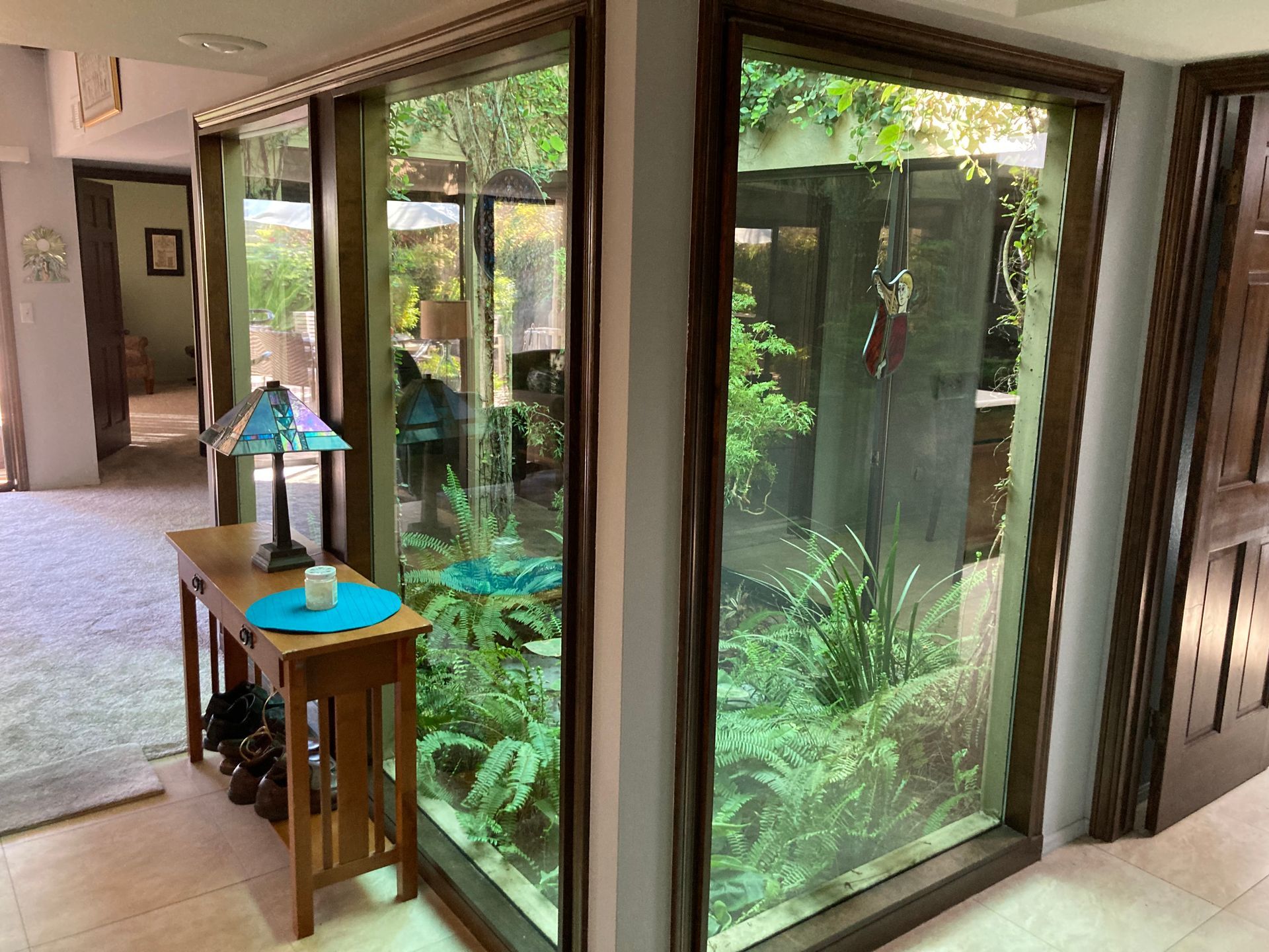 A small wooden table stands beside large glass windows looking into a green, plant-filled indoor courtyard.