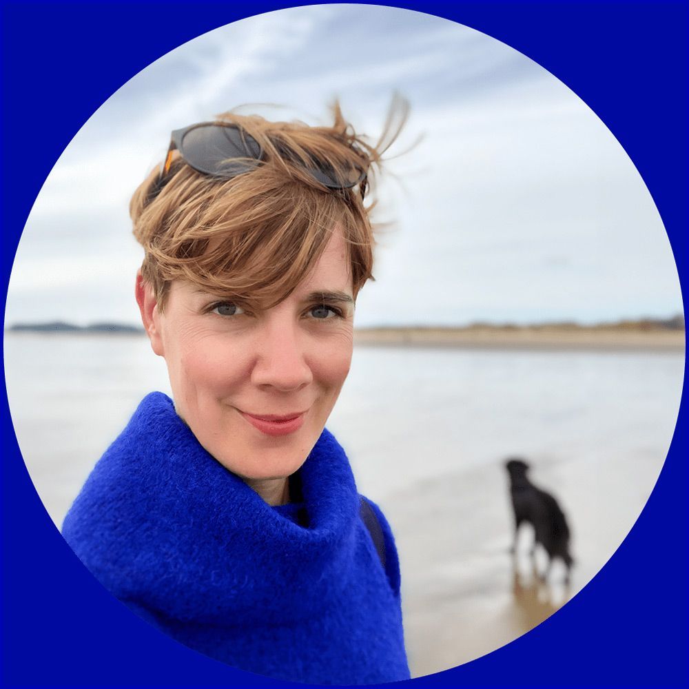 Tonya mit einem blauen Wollpulli steht an einem Strand, im Hintergrund ist ihre Hündin Yani zu sehen.
