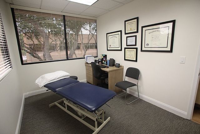 A doctor 's office with a bed , desk , chair and certificates on the wall.