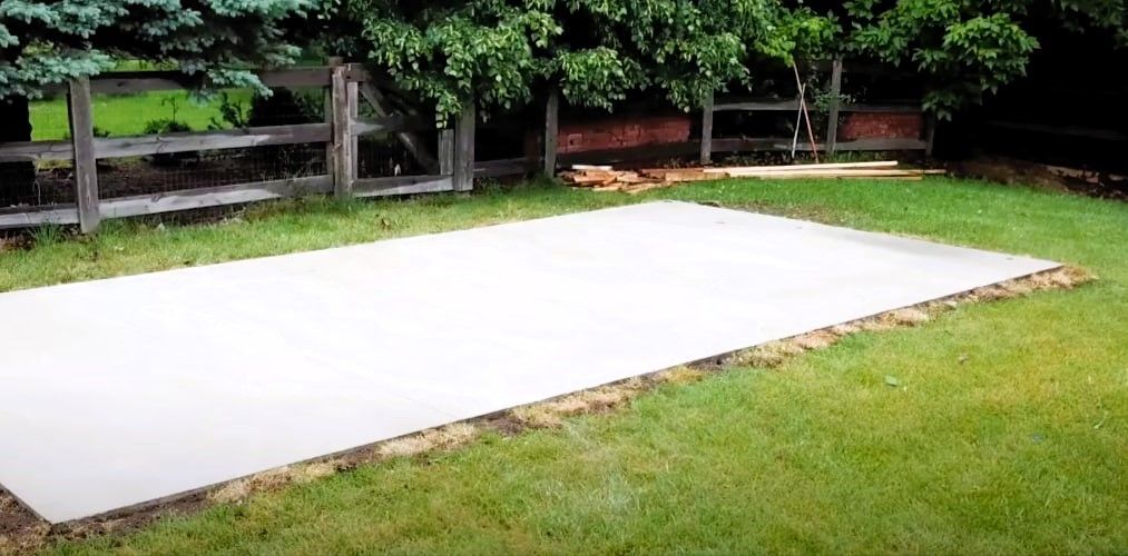 Freshly poured concrete slab in a grassy backyard with a wooden fence and trees.