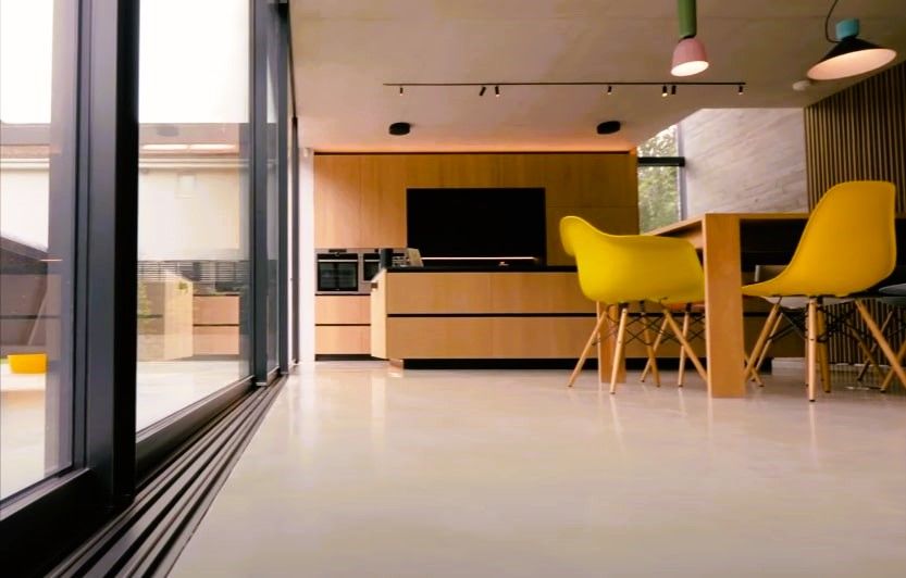 Modern kitchen with large windows, wooden cabinets, yellow chairs, pendant lights, and polished concrete floor.