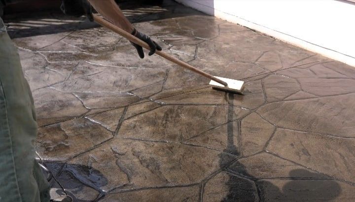 Person sealing or cleaning stamped concrete surface with a brush in a sunny outdoor setting