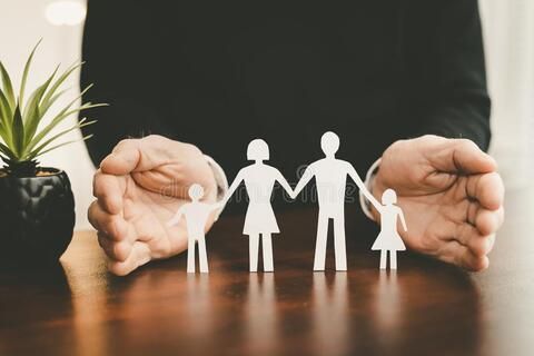 Hands protect a paper cutout family on a wooden table, next to a small plant.