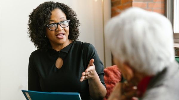 Woman in glasses gestures while speaking to older person in an indoor setting.