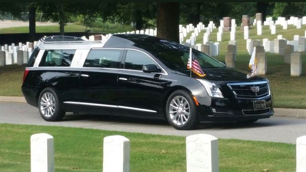 Black hearse with American flag drives past gravestones in a cemetery.