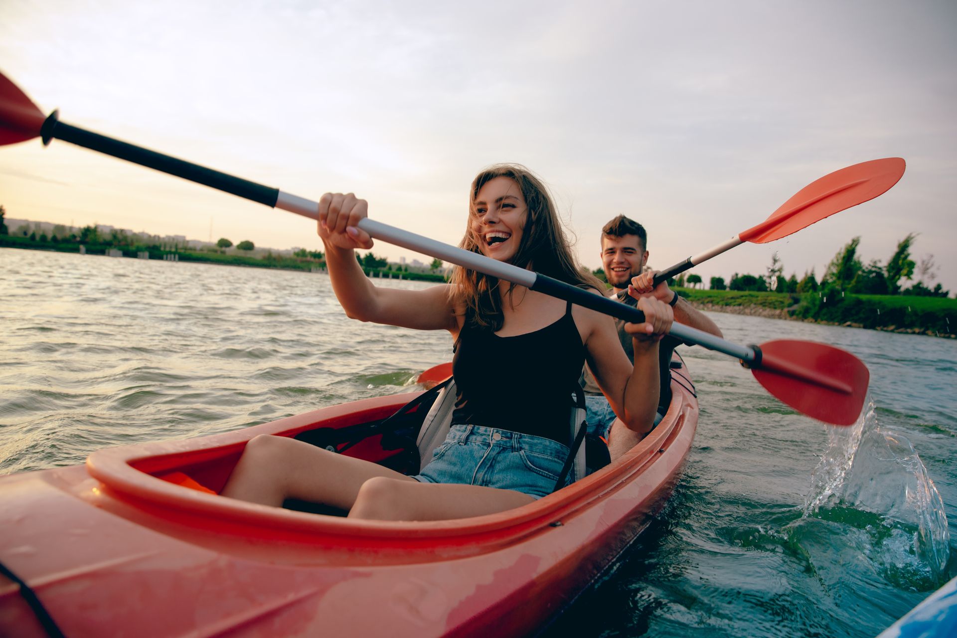 Woman and man laughing while kayaking on a lake with red kayak, blue water, and paddles.