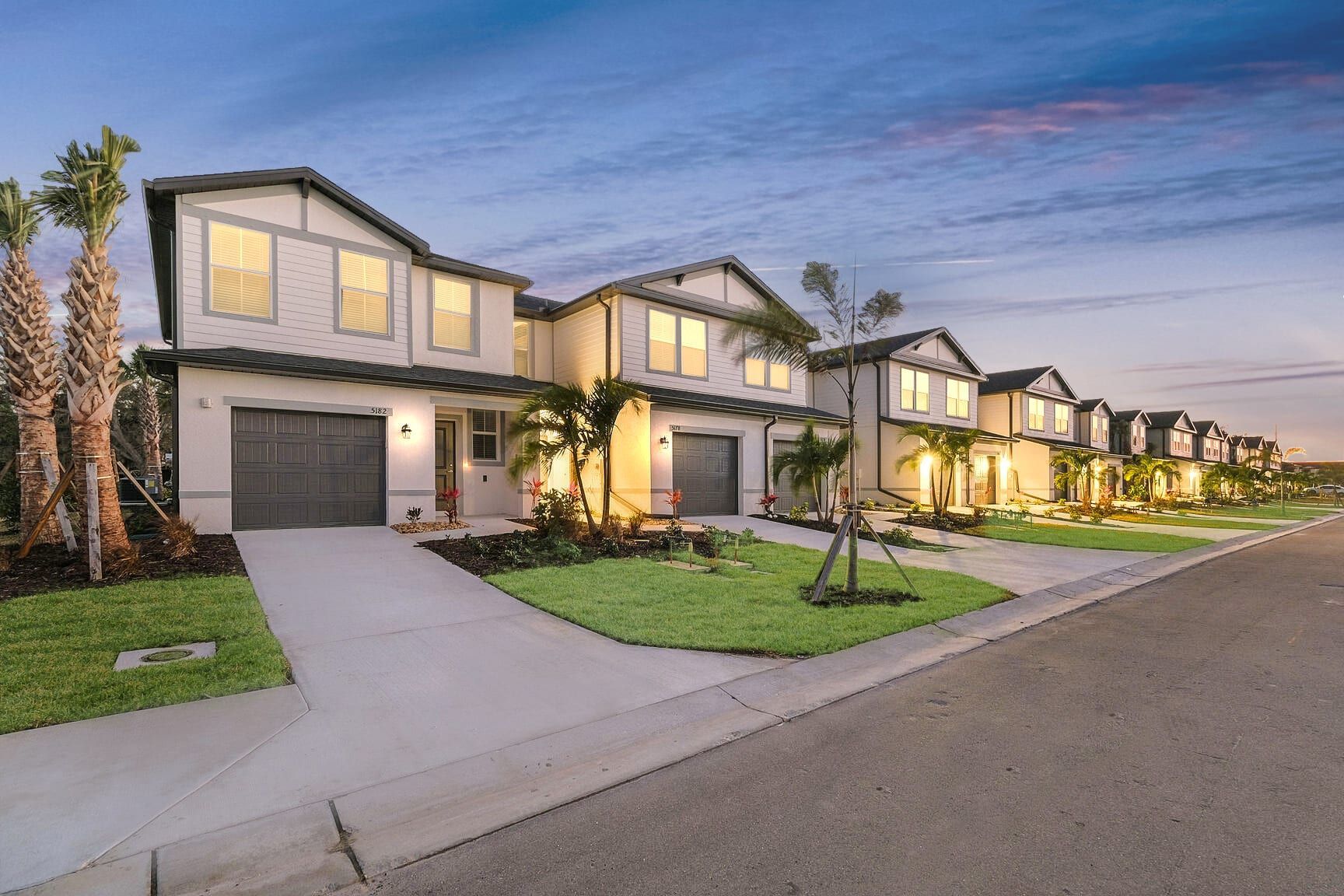 A row of modern, light-colored townhouses with garages at dusk, lined with palm trees and a paved street.