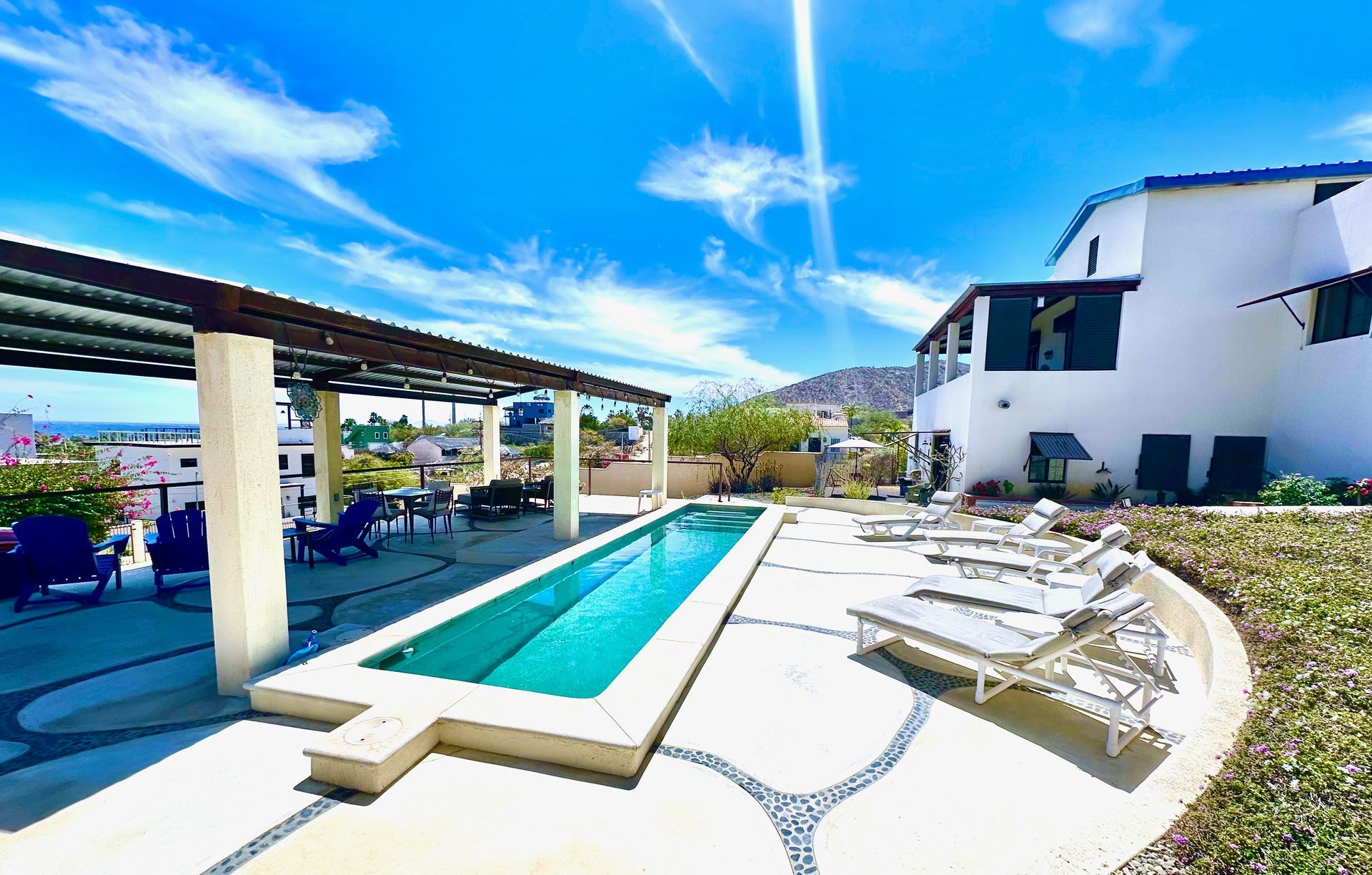 Swimming pool, lounge chairs, and pergola on a patio with a building and blue sky in the background.