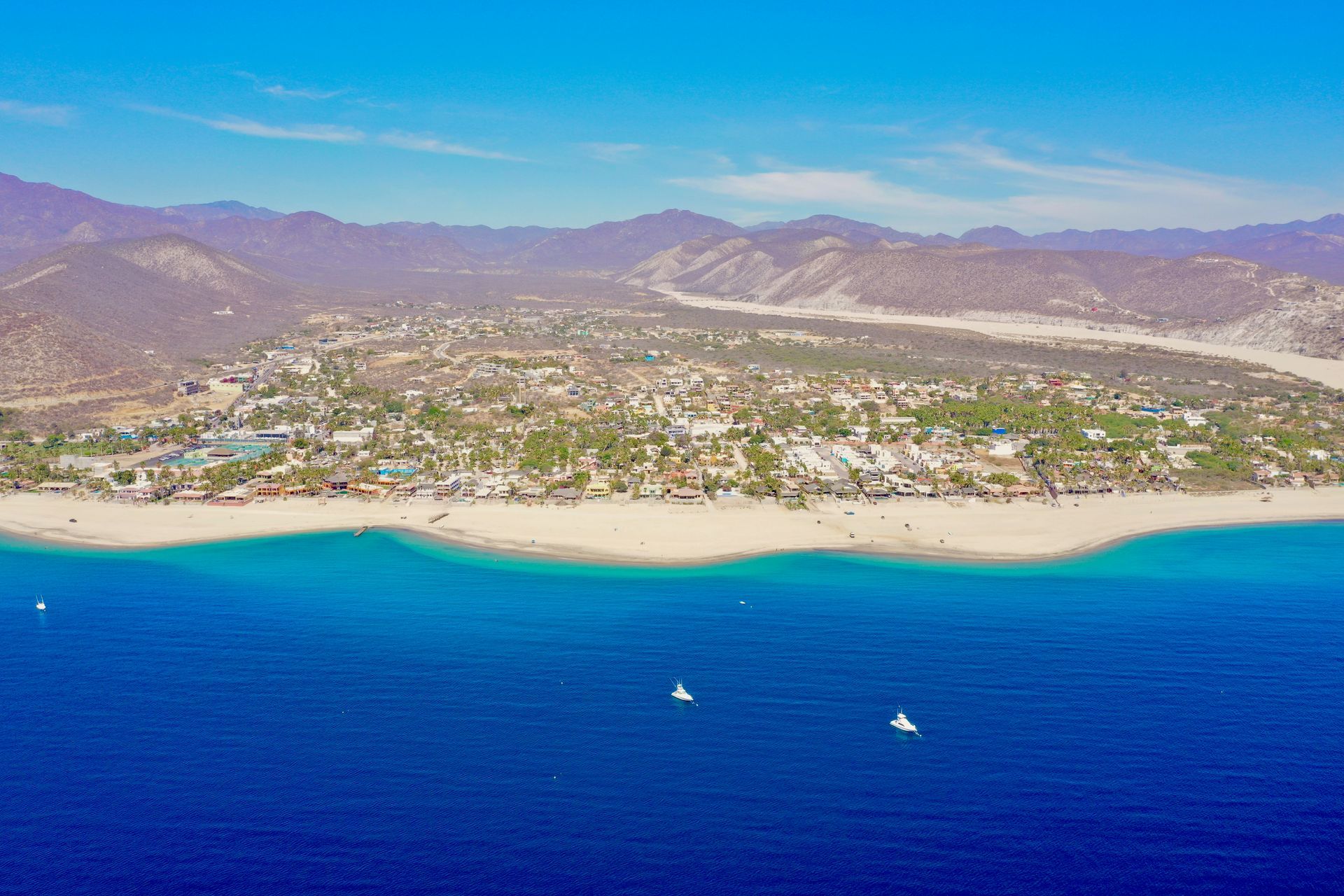 Aerial view of a coastal town with sandy beach, turquoise water, and mountains in the background