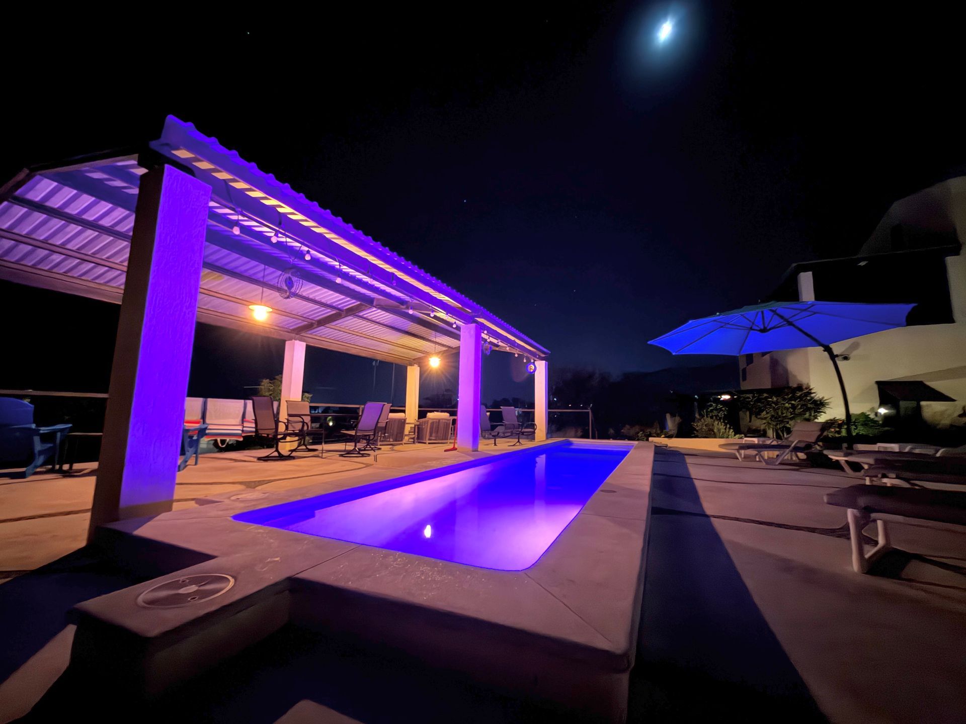 Nighttime view of a purple-lit swimming pool under a covered patio; umbrella, furniture, and moon visible.