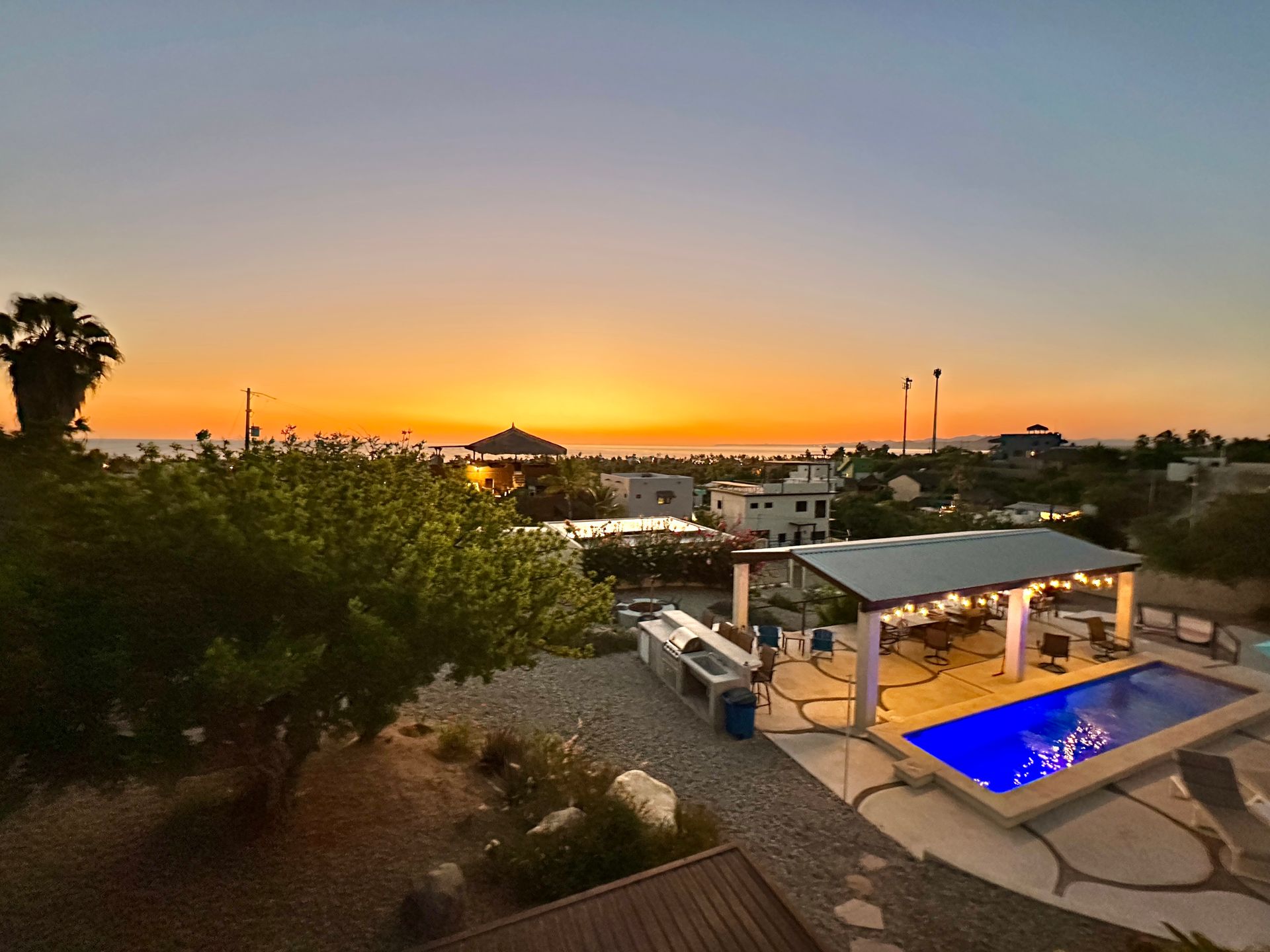 Sunset view over a pool and outdoor kitchen with a blue-lit pool against an orange sky.