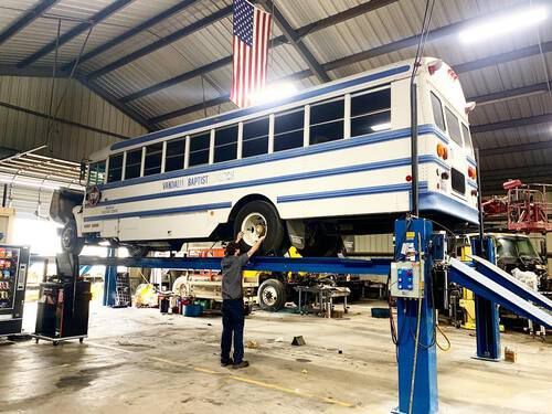 A man is standing next to a bus on a lift in a garage.