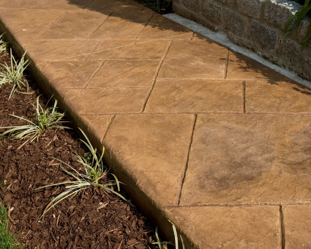stamped concrete sidewalk stained red-brown with dirt and sprouting plants along the side