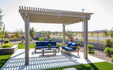Square concrete slab sits in the middle of a grass patch with a ramada and blue patio furniture sitting on top