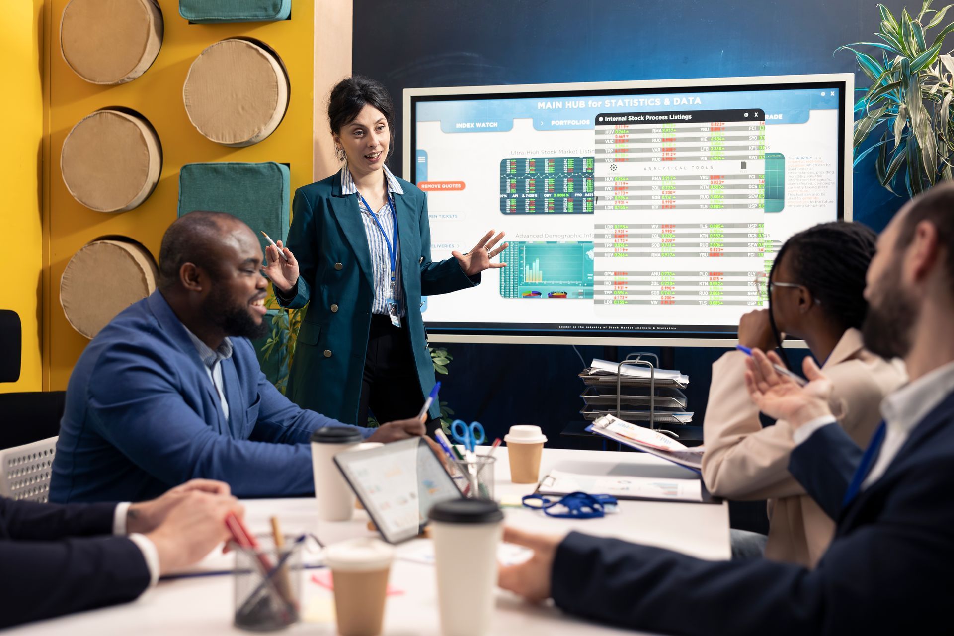 A presenter leads a professional team in a meeting, gesturing toward a large screen displaying complex data charts.