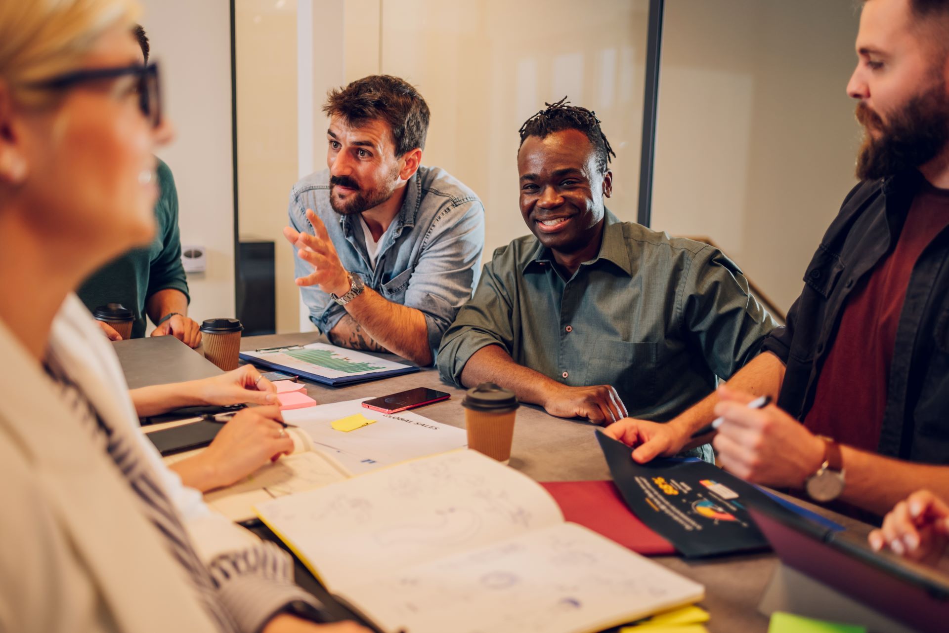 People in a business meeting, gathered around a table; discussing ideas and looking at papers, smiling.