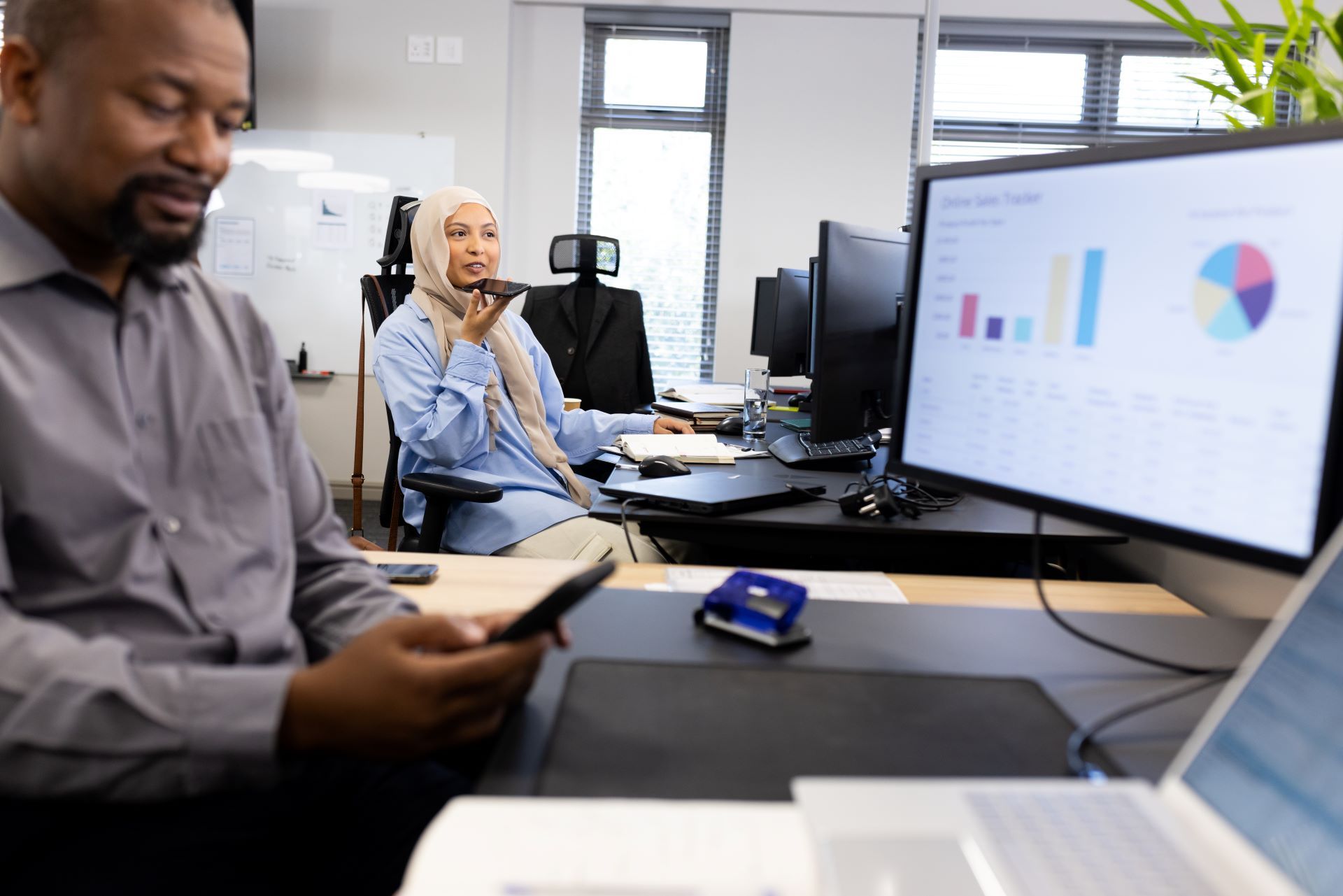 Office setting: Man using phone, woman on call near monitor with graphs.