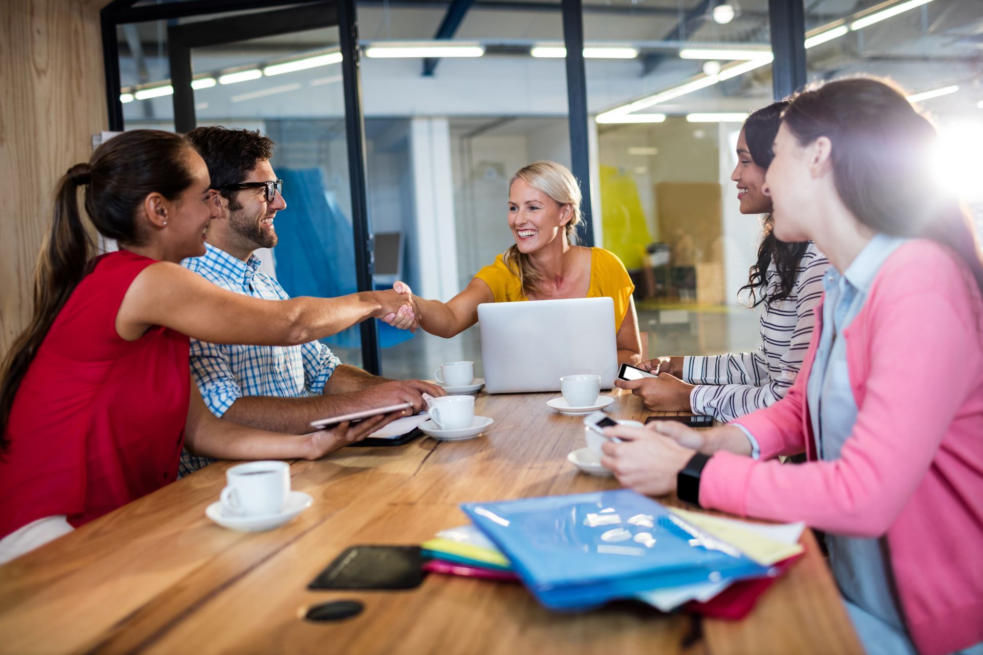 A business meeting in an office setting. Two people shaking hands over a wooden table, others are watching and smiling.
