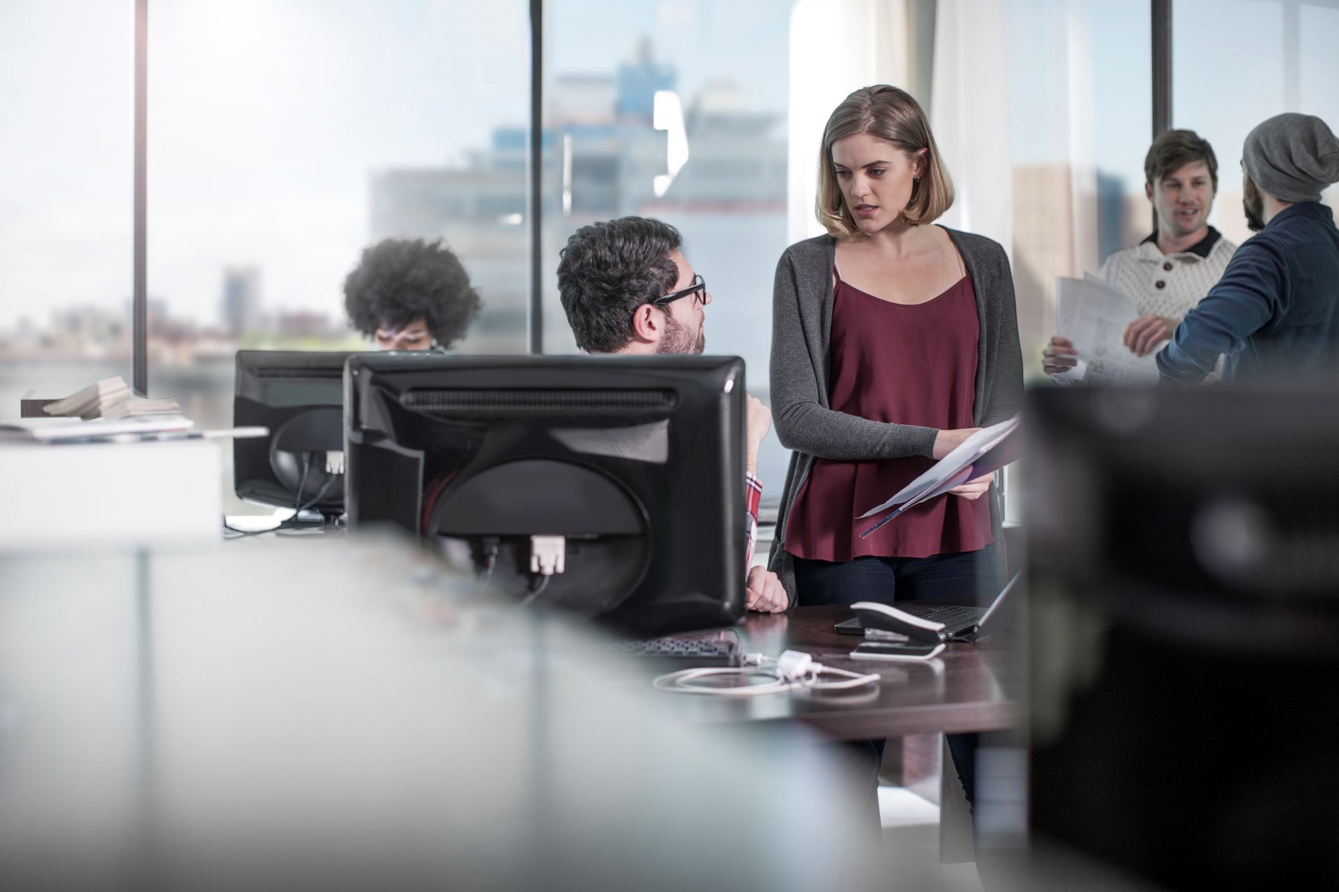 A woman is standing in front of a computer in an office talking to a man.